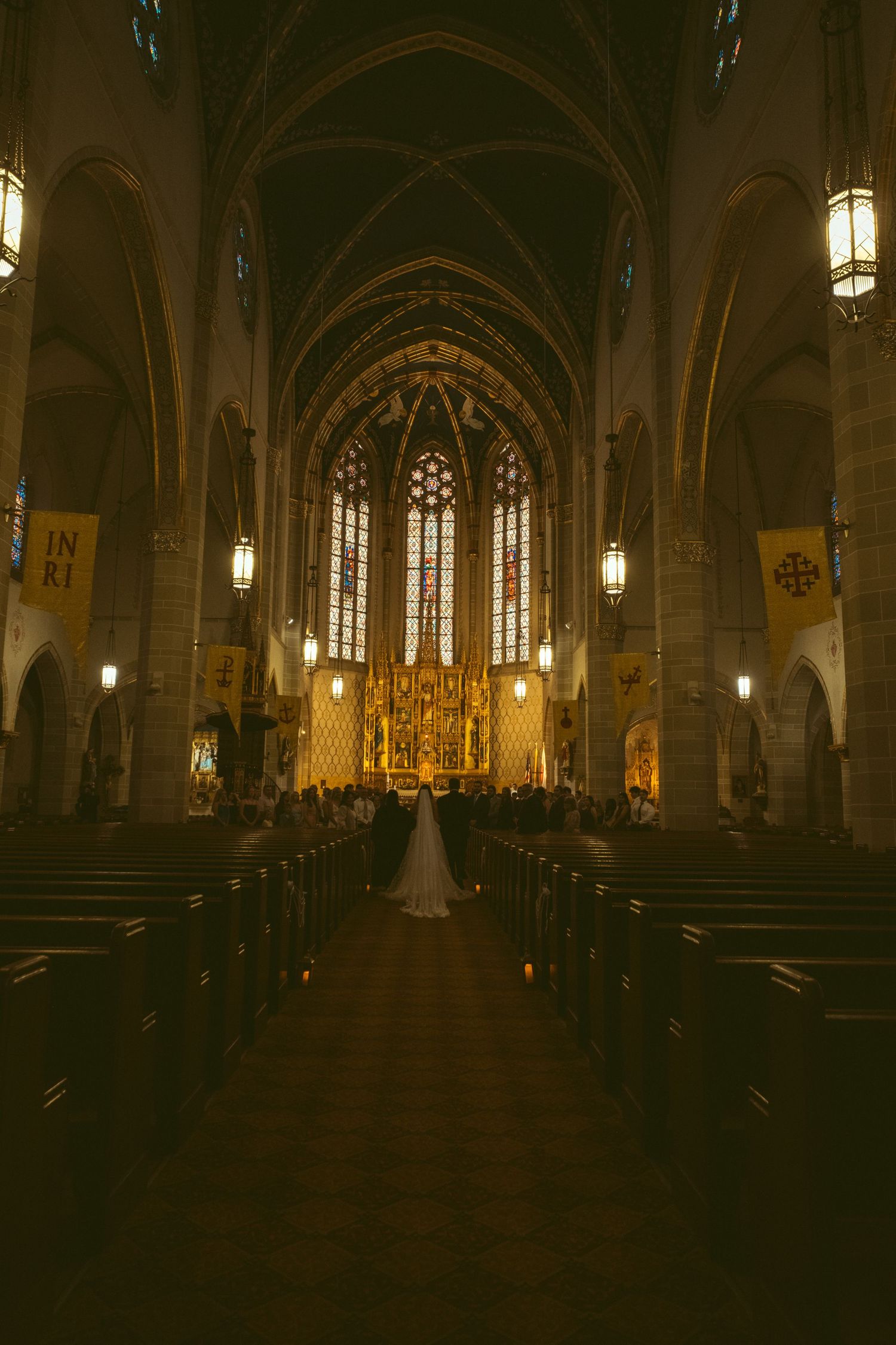Bride walks down the aisle of a grand Gothic cathedral with stained glass windows and golden altar during wedding ceremony.
