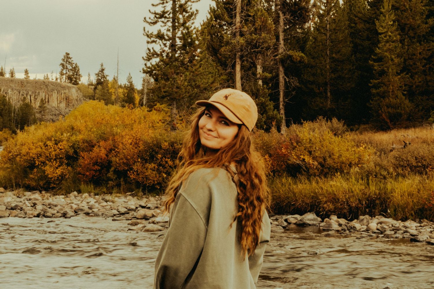 Person in a baseball cap and sweater smiles by a river with autumn foliage and evergreen trees in the background.