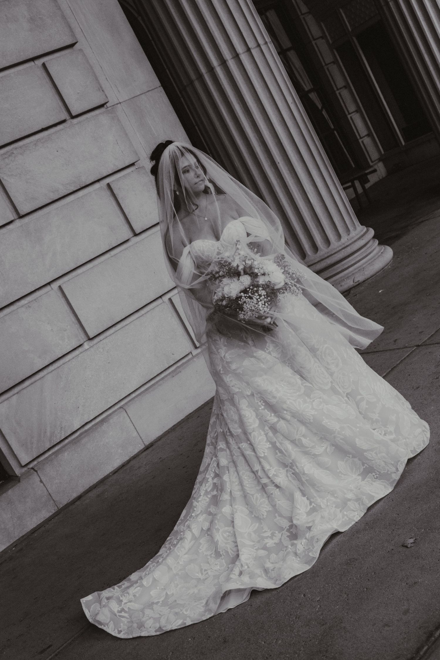 A bride in a flowing lace wedding gown poses gracefully in front of a classic stone building with columns.