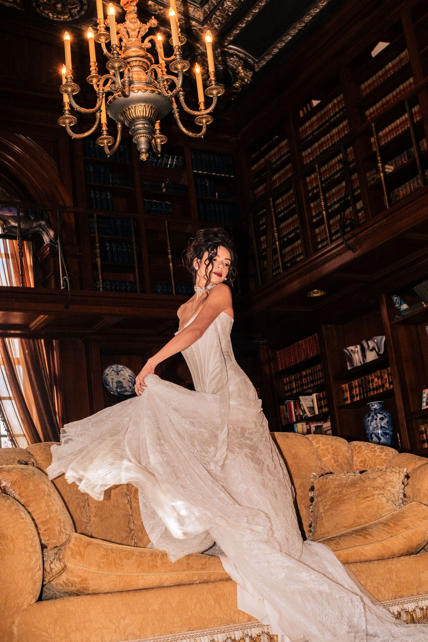 A bride in a flowing white gown lounges elegantly on a vintage couch in a grand library with chandeliers overhead.