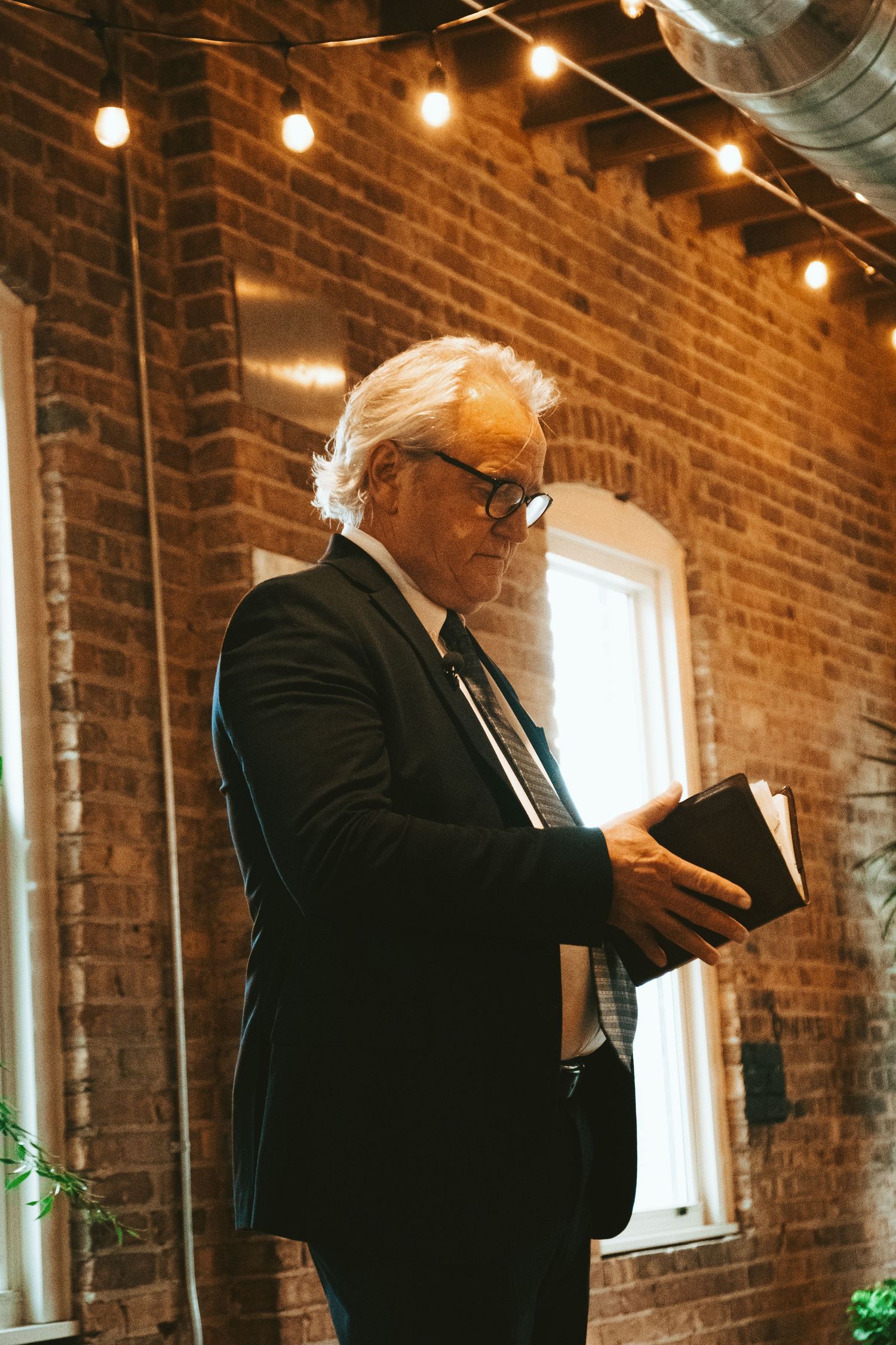 Elderly man in suit reading book against brick wall with string lights.