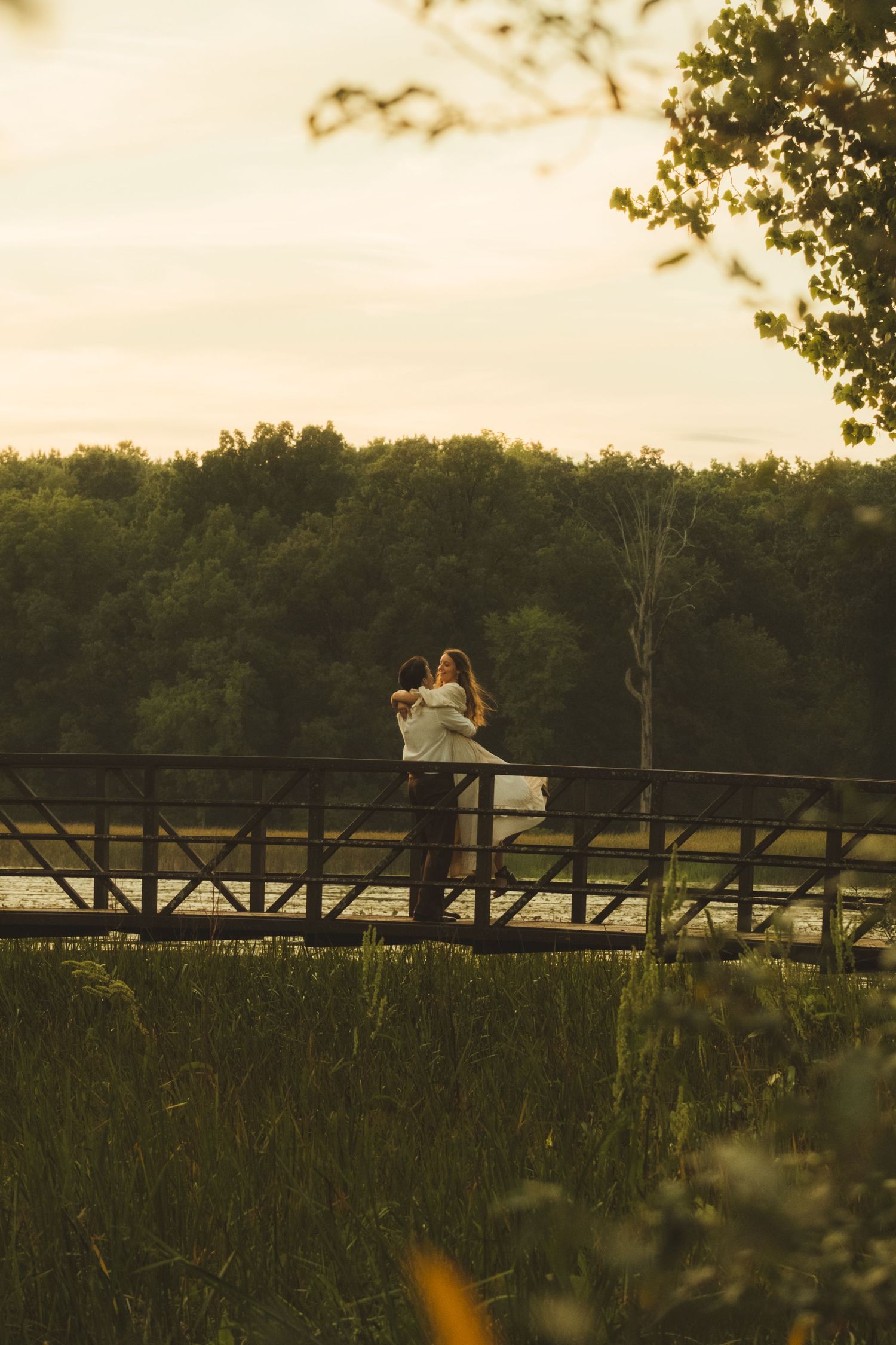 Couple embracing on a rustic wooden bridge surrounded by lush green trees at golden hour sunset.