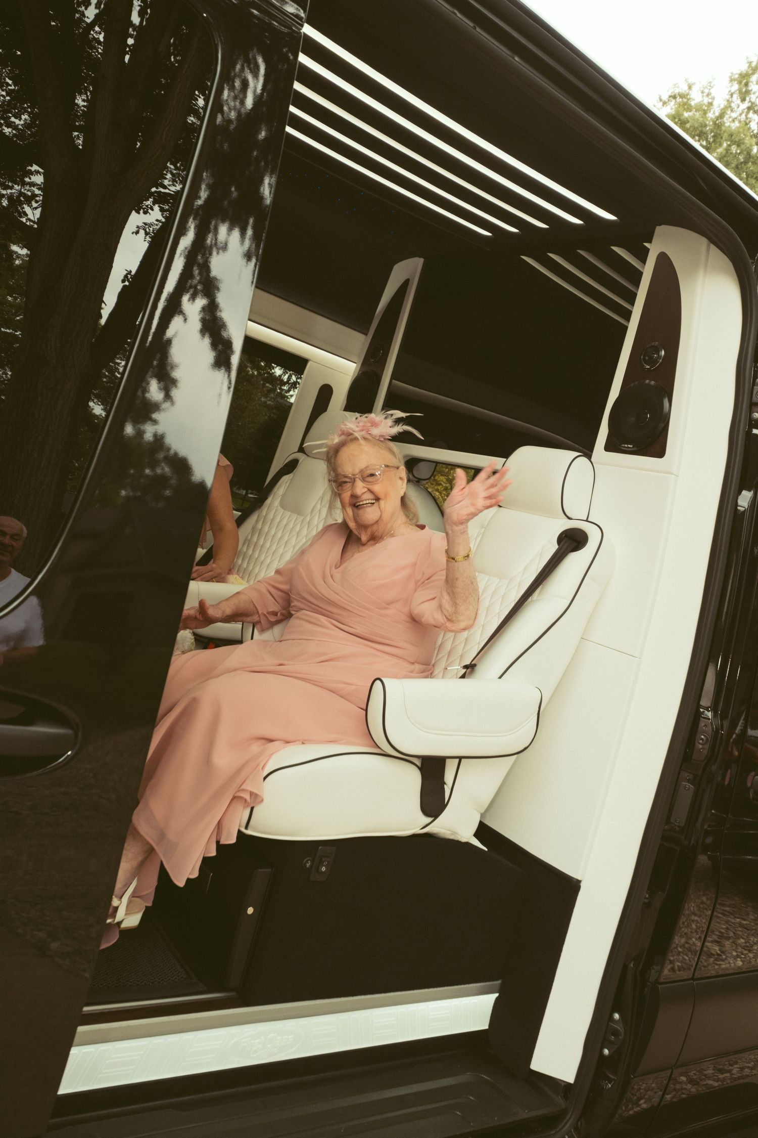 Elderly woman in pink dress waving happily from luxury white limousine interior seat.