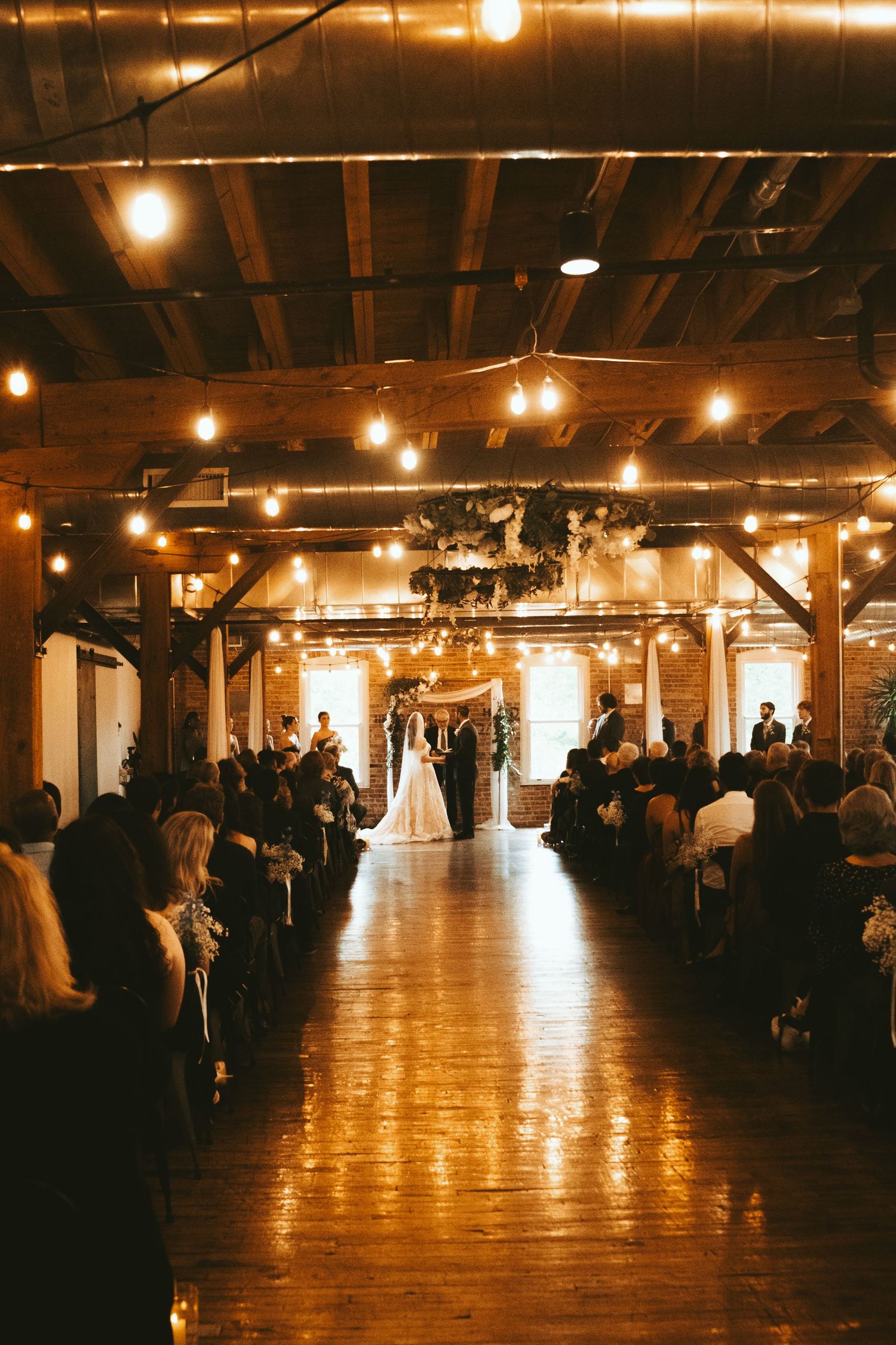 A wedding ceremony in a rustic industrial venue with exposed brick walls and warm string lighting overhead.
