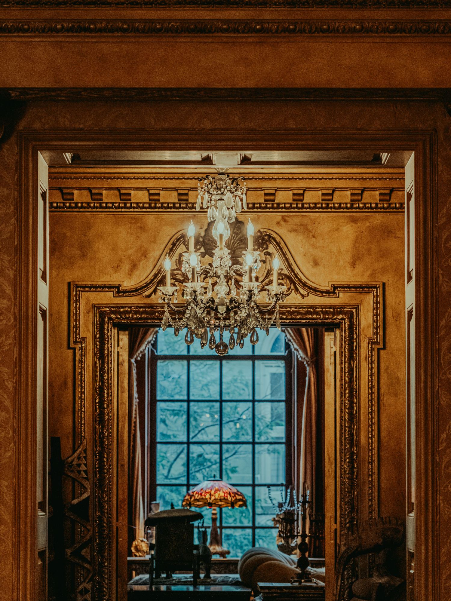 Ornate golden room with crystal chandelier, decorative molding, and elegant window view in a luxurious vintage interior.