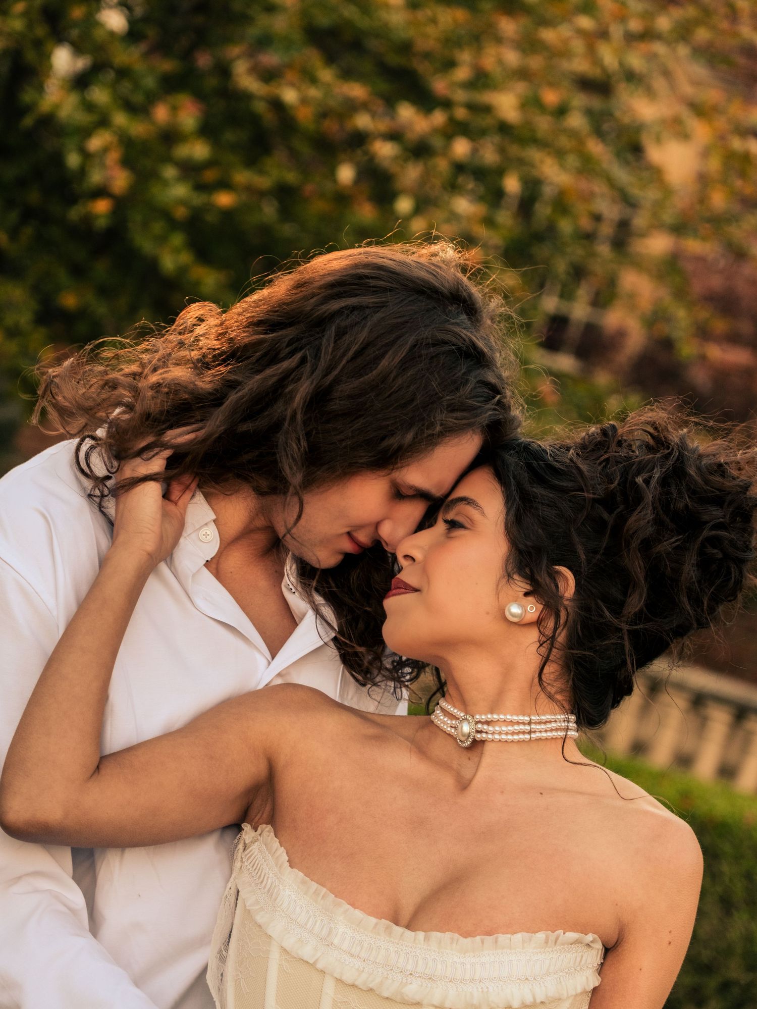 A romantic couple in wedding attire share an intimate moment outdoors during golden hour with autumn foliage in background.