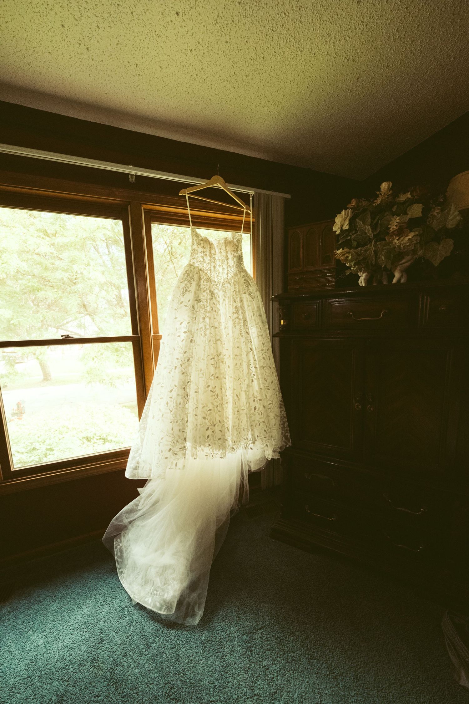 A elegant white lace wedding dress hanging on a wooden hanger in front of a sunlit window in a cozy room.