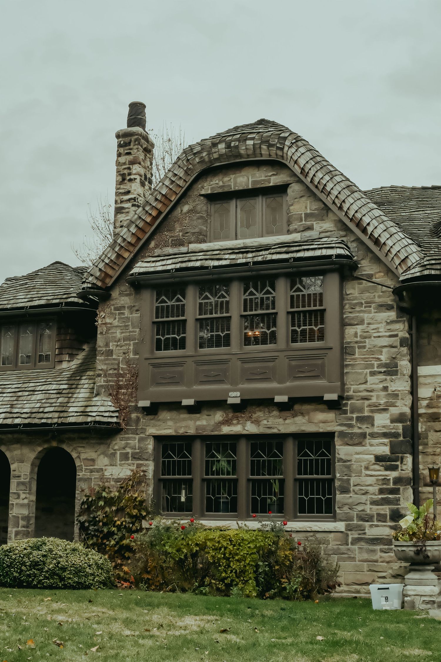Historic Tudor-style stone mansion with arched windows, curved gabled roof, and brick chimney on overcast day.