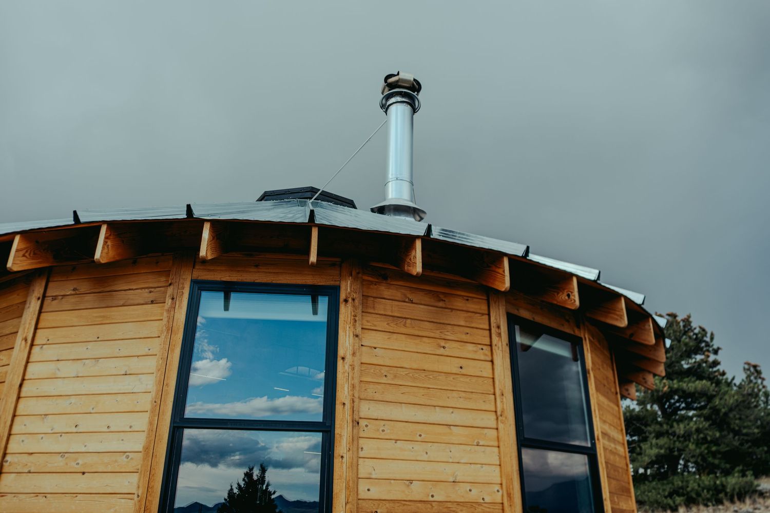 Wooden circular cabin with metal chimney pipe on curved roof against overcast sky.