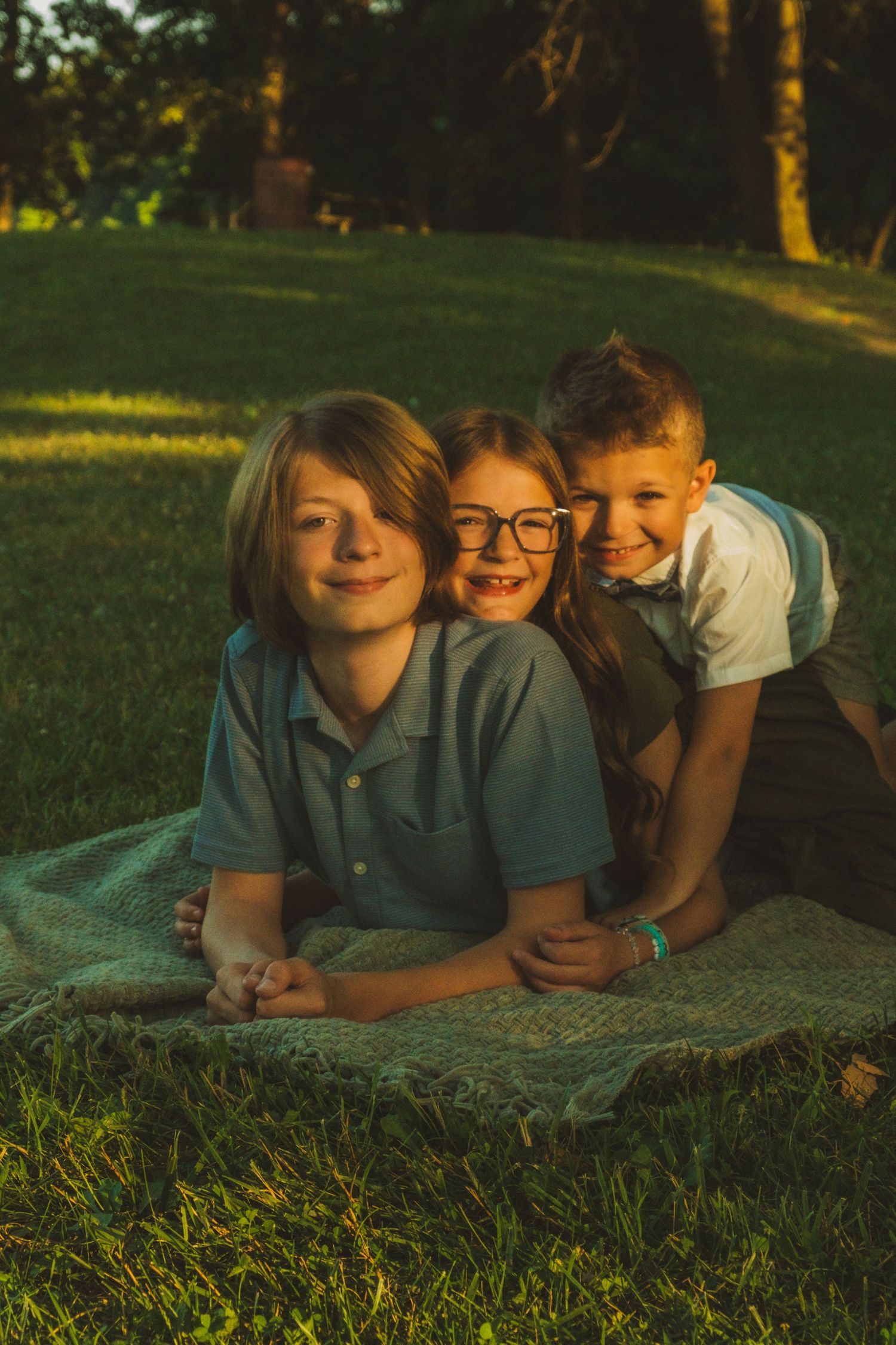 Three children lie together on a blanket in the grass during golden hour sunlight.