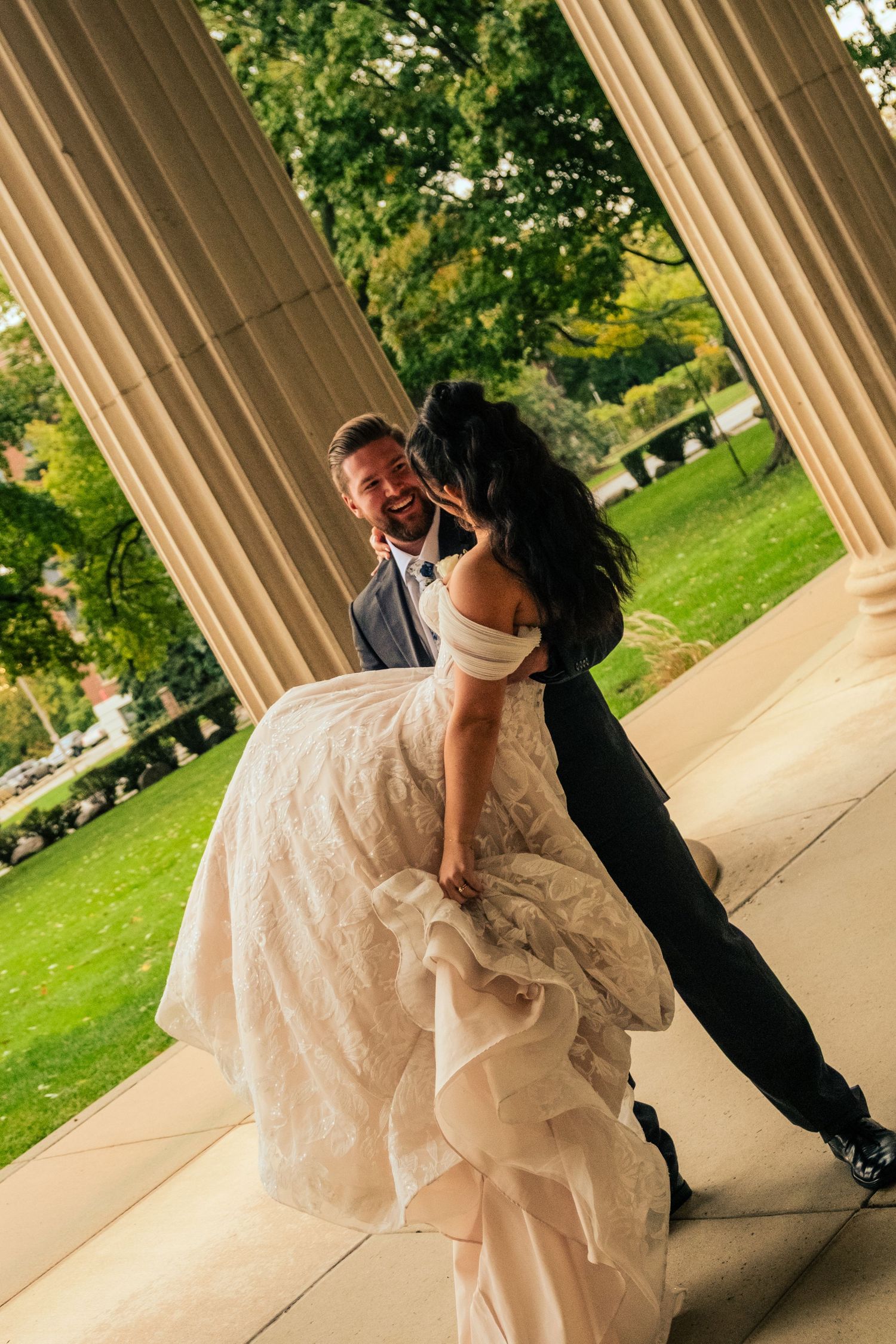 A couple in wedding attire share a romantic moment between elegant stone columns on a sunny day in a garden setting.