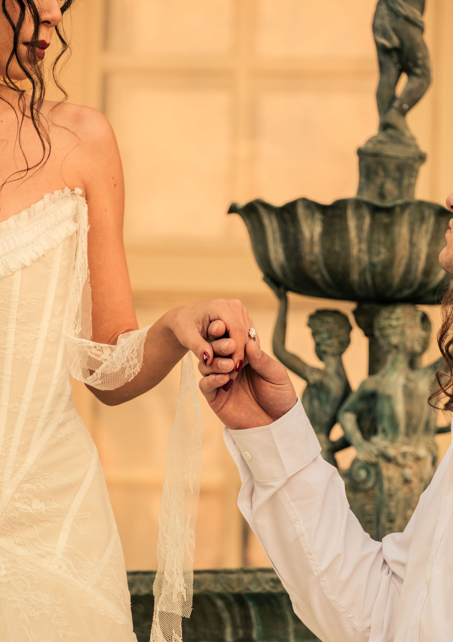 A couple holds hands wearing white formal attire against a warm peach-toned background with ornate sculptures.