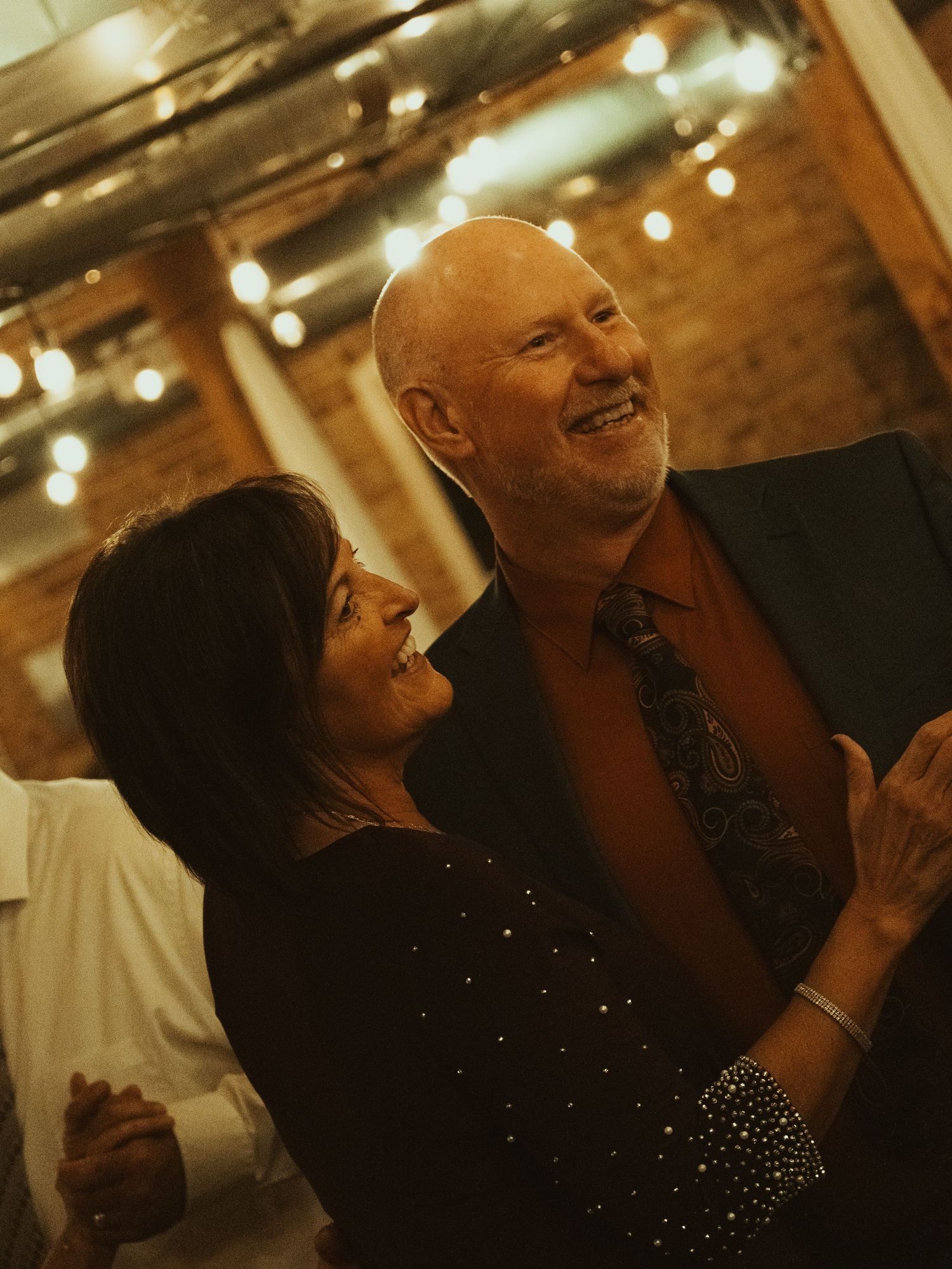 Two people share a joyful moment dancing together in a warmly lit venue with string lights overhead.