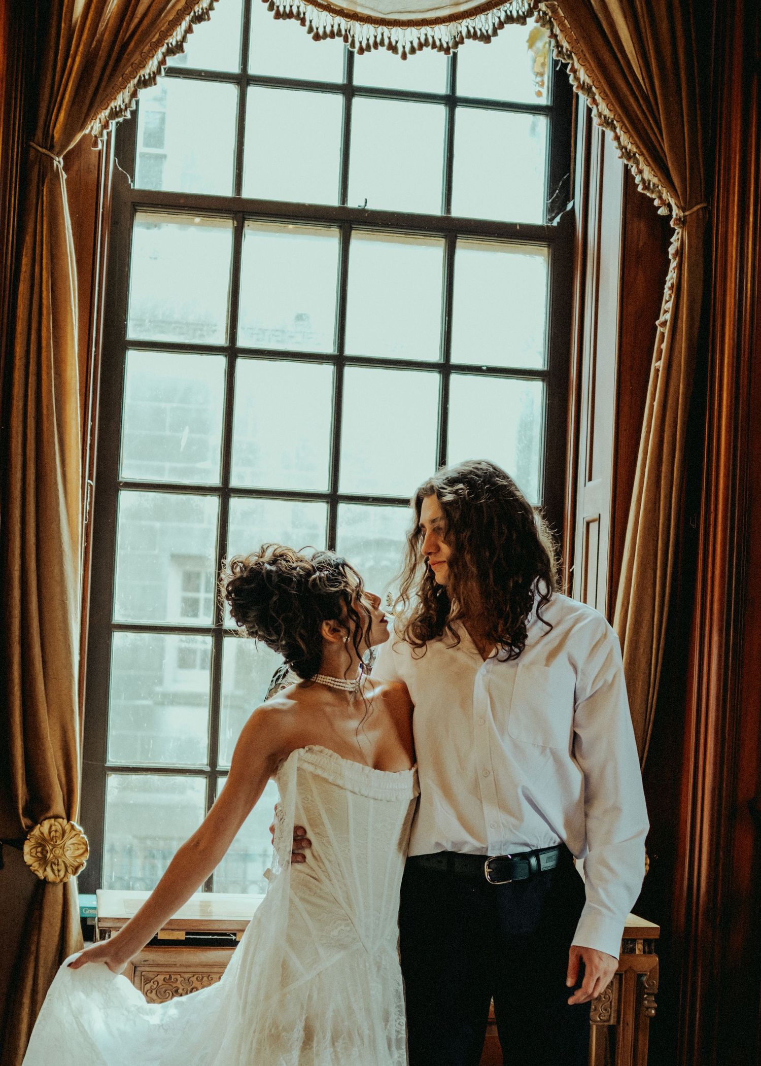 Wedding party members in formal attire look out large windows in an elegant room with brown curtains and city views.