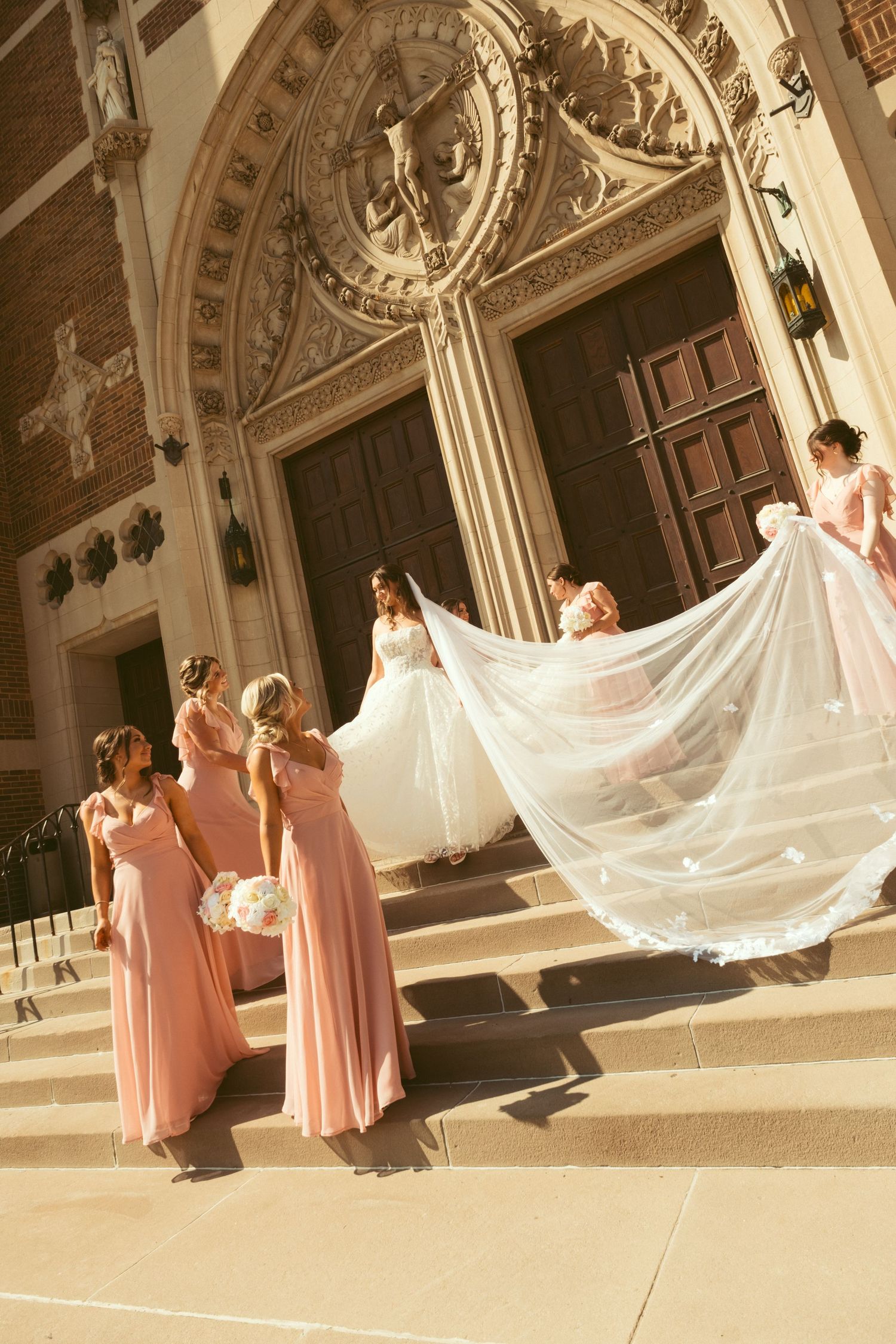 Bride in white gown with bridesmaids in pink dresses holding veil on church steps.