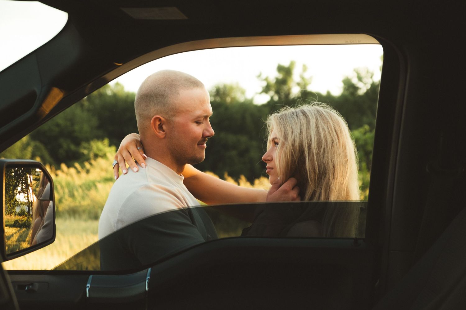 Couple sharing a romantic moment outside a car window in a sunny field at golden hour.