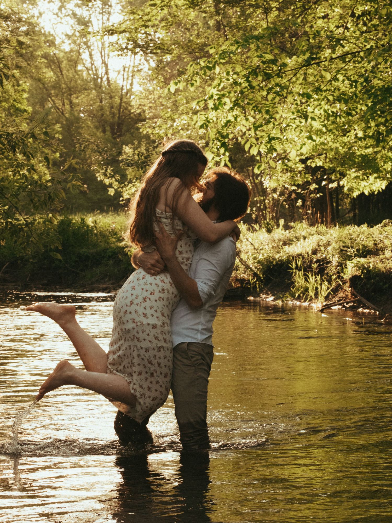 A couple sharing a romantic kiss while standing in a shallow sunlit river surrounded by lush green trees.