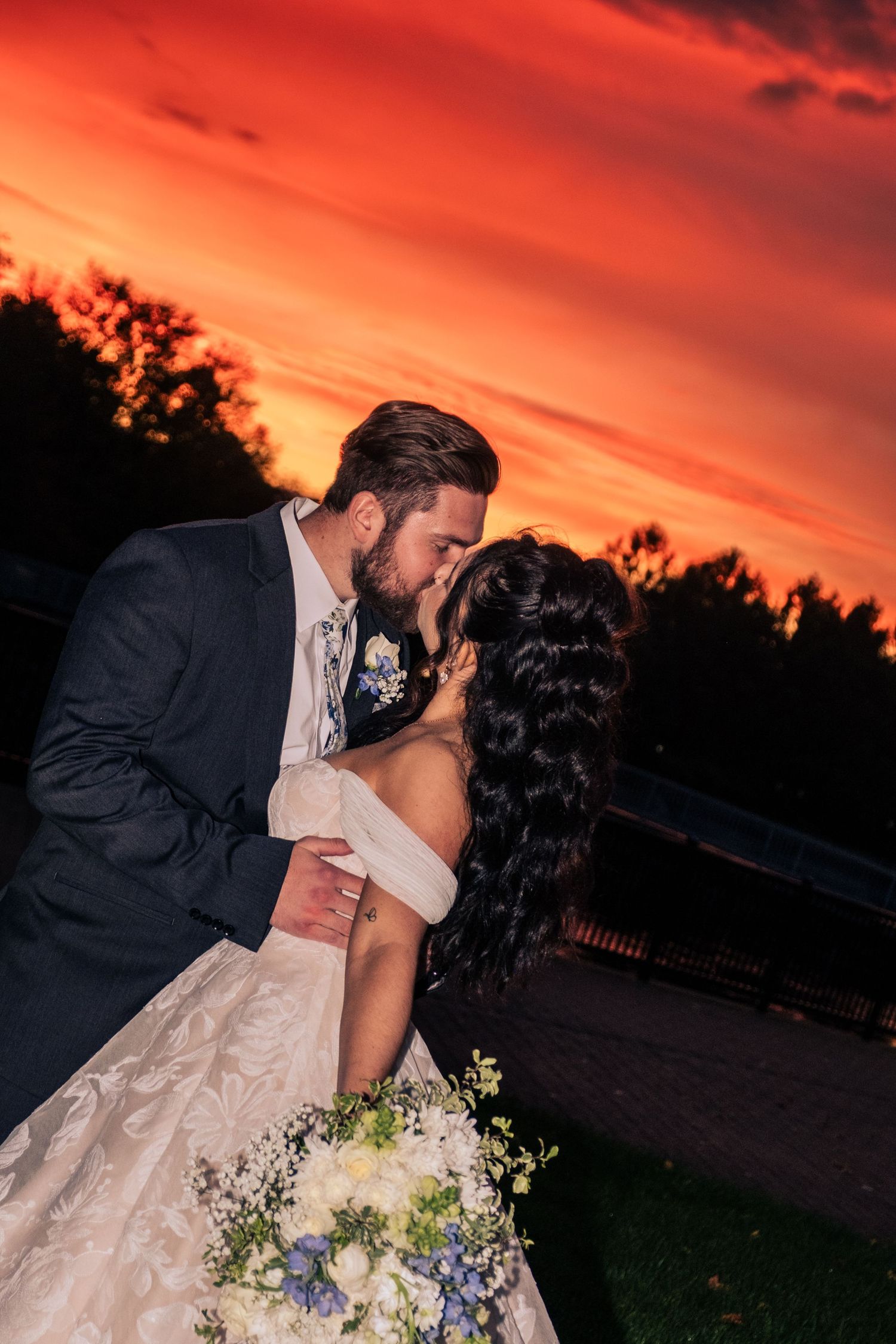 A romantic wedding couple shares a kiss against a vibrant orange and red sunset sky.