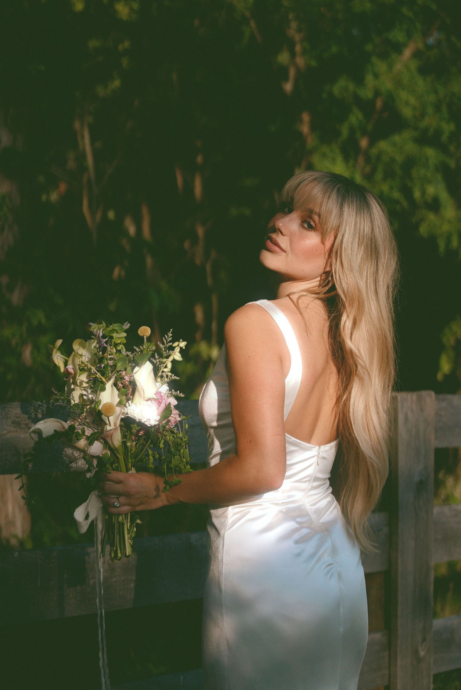Woman in white dress holds delicate floral bouquet while posing gracefully against lush green garden backdrop at sunset.