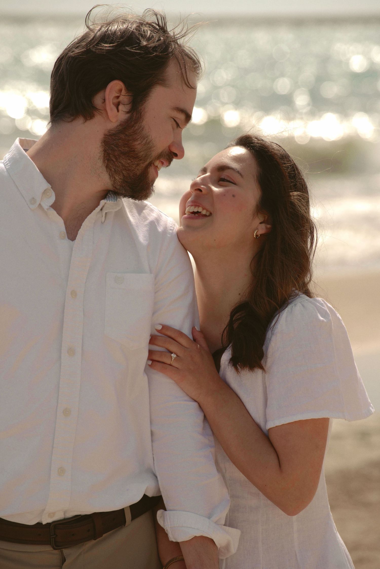 A couple in white attire shares a joyful intimate moment together on a sunlit beach.