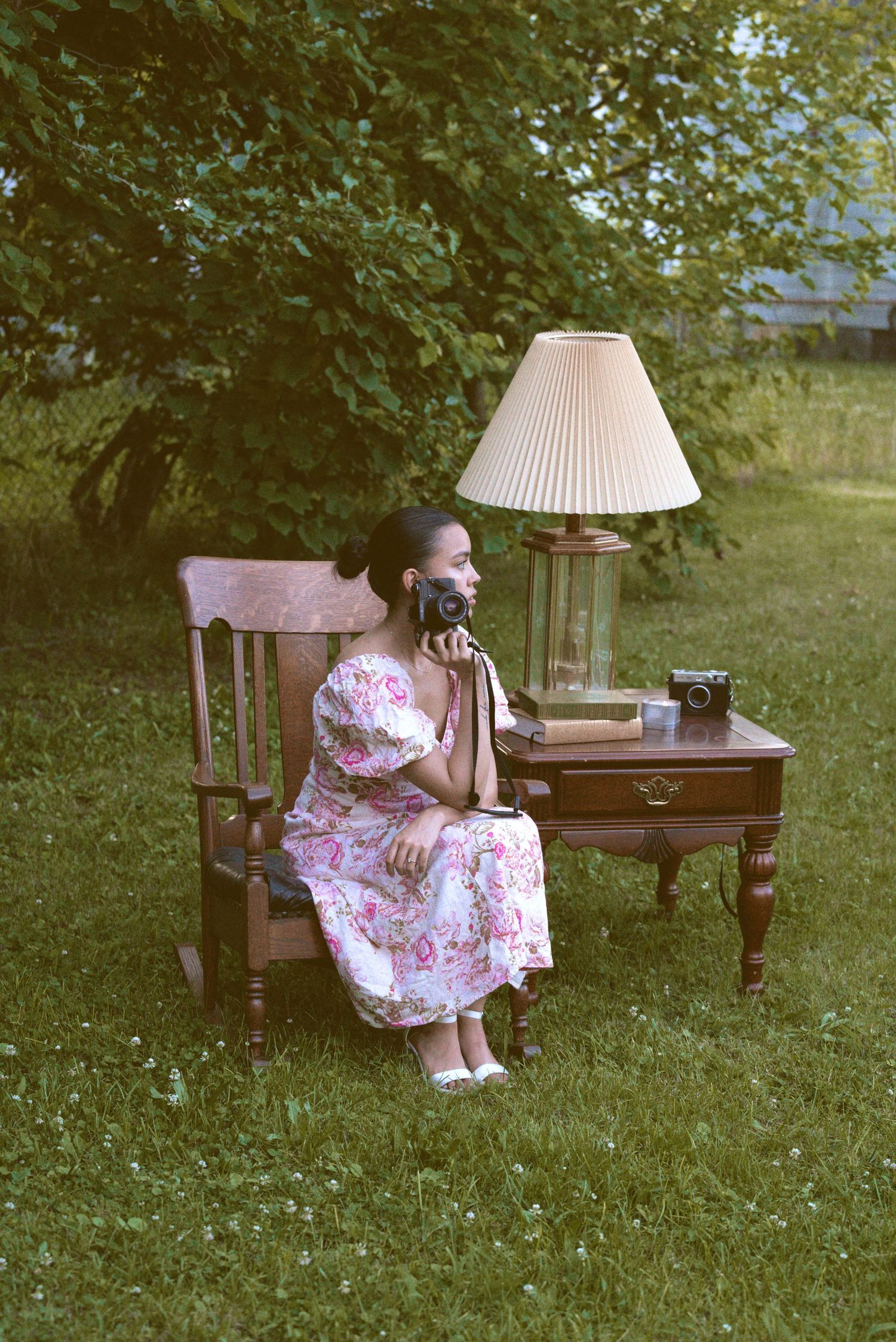 Vintage scene on a lawn with wooden chair, side table with lamp, and person in floral dress sitting outdoors.