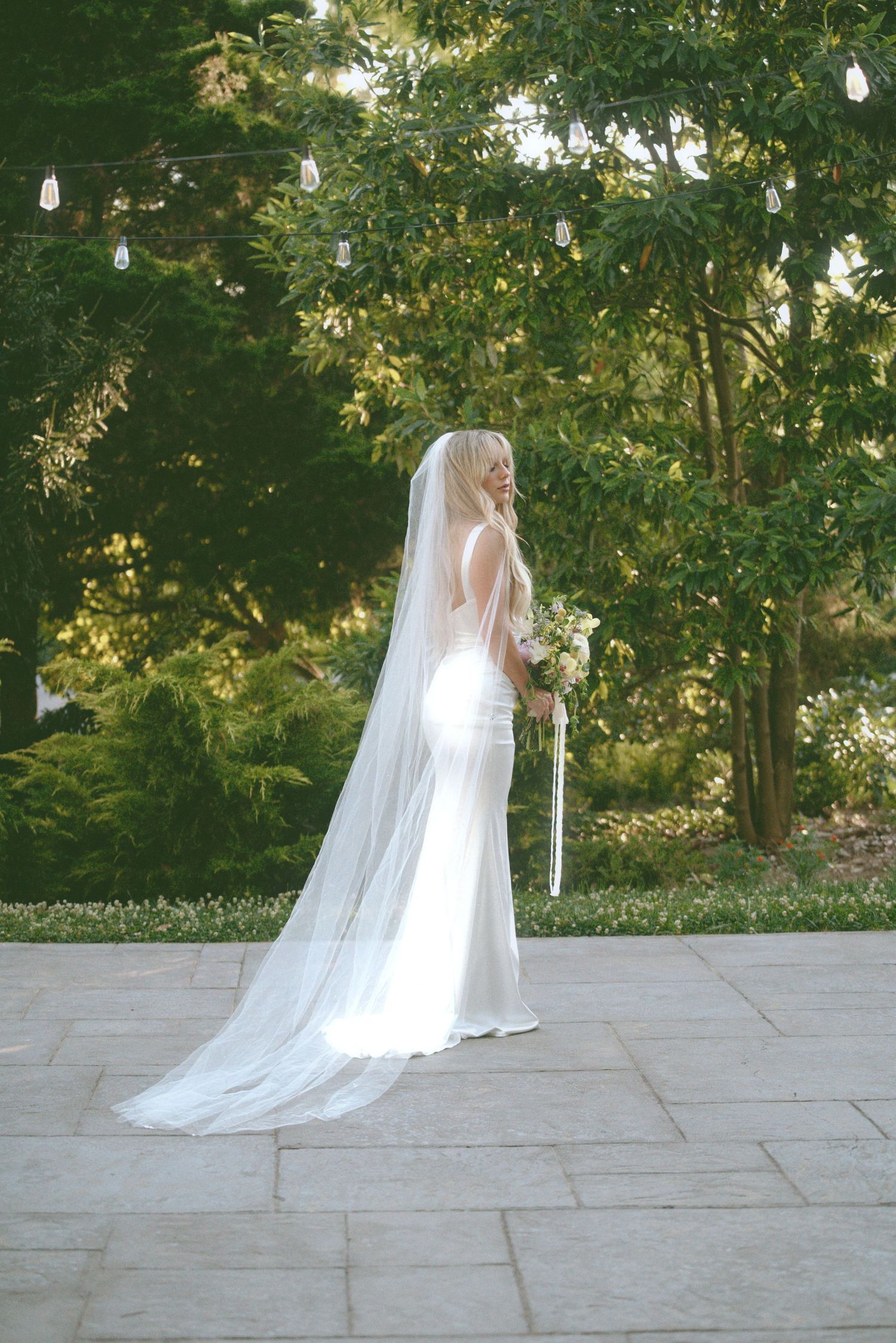A bride in a flowing white gown and long veil stands on stone paving surrounded by lush garden greenery and hanging lights.