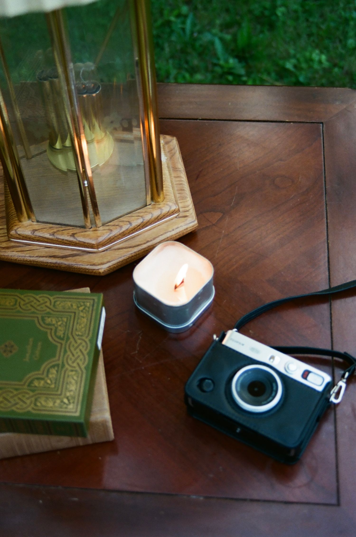 A vintage instant camera sits on a wooden table alongside old books, a lantern, and a small glowing candle.