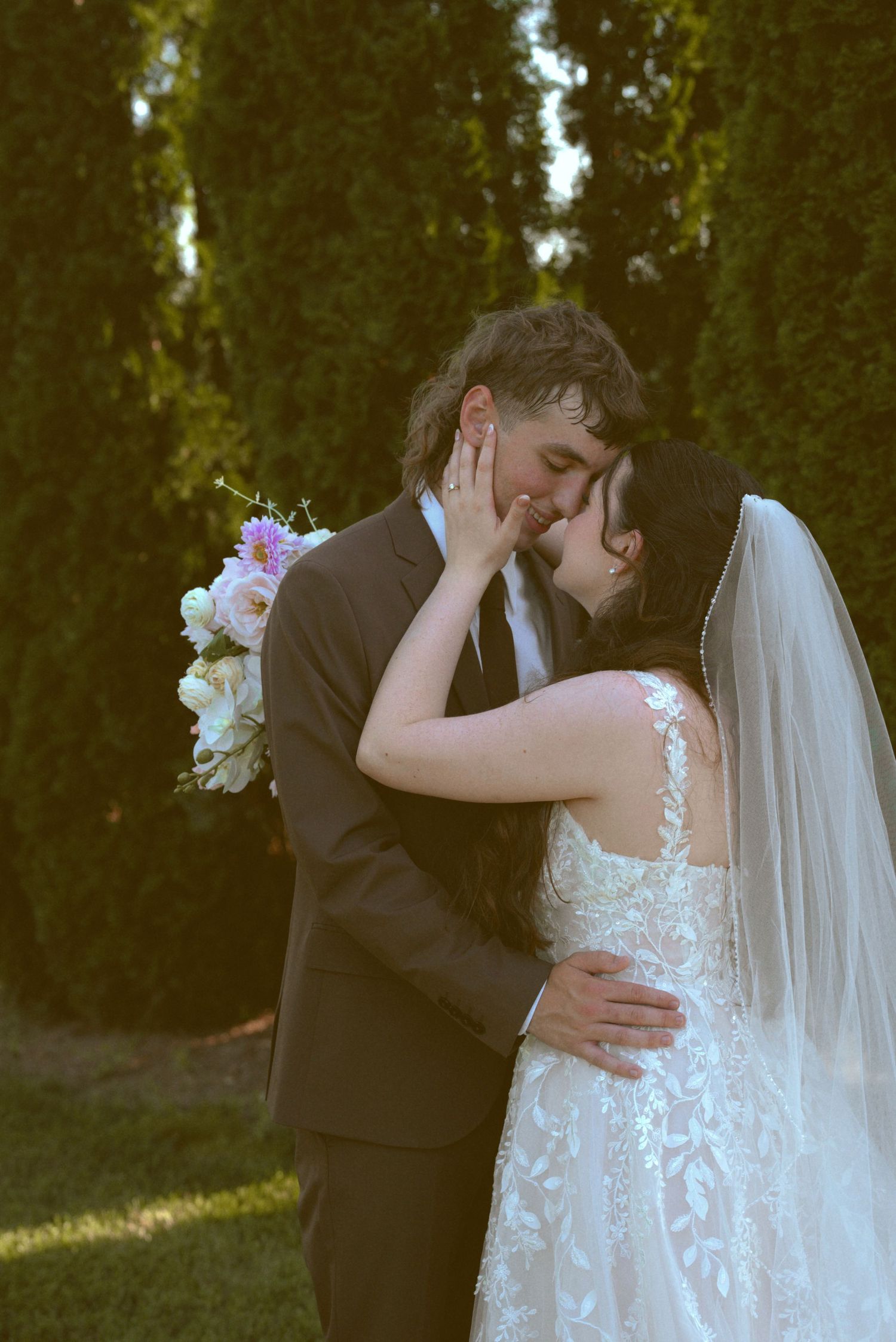 Romantic wedding couple embraces during sunset in garden with evergreen trees and floral bouquet.