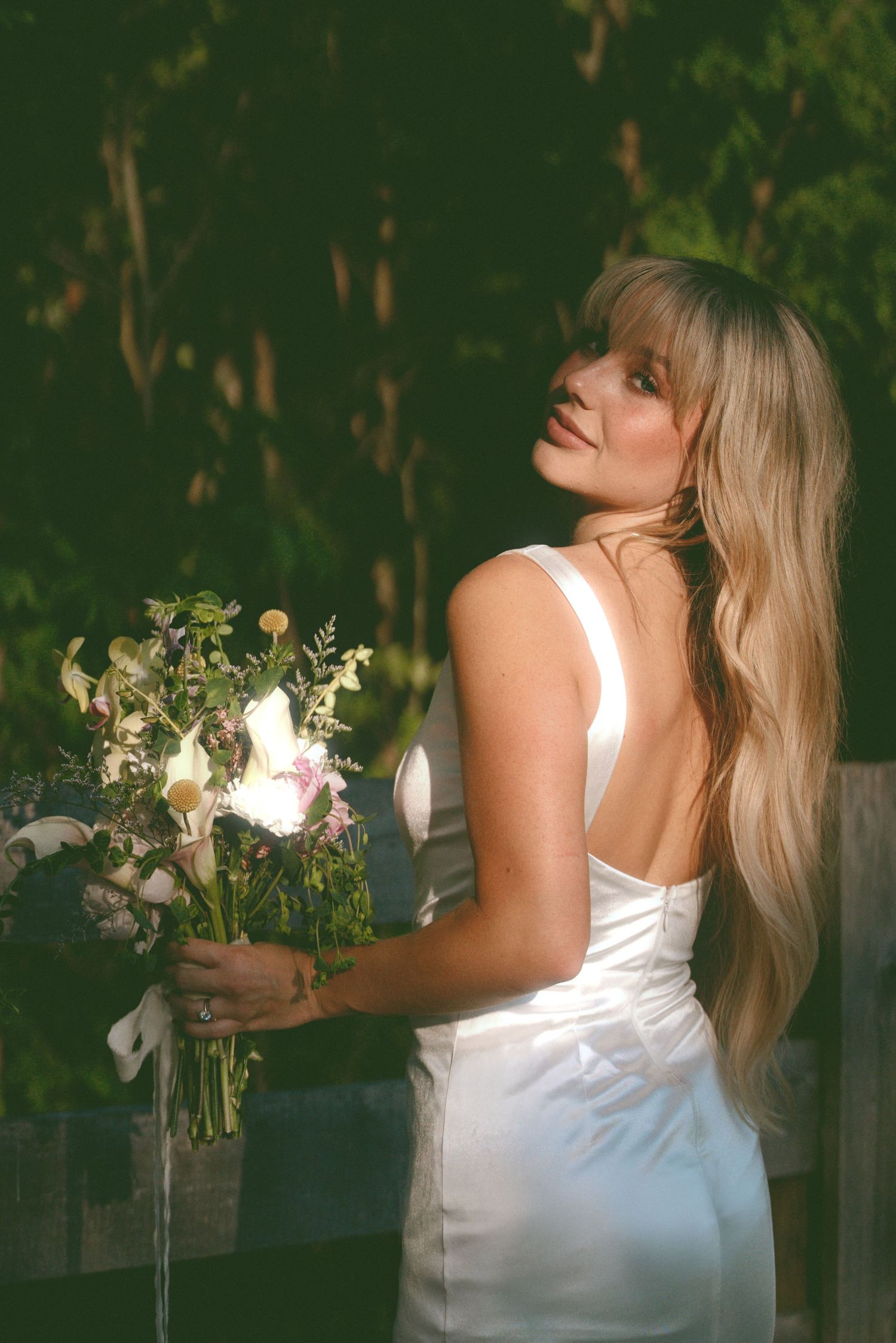 A figure wearing a white dress holds a bouquet of flowers in an outdoor garden setting at dusk.