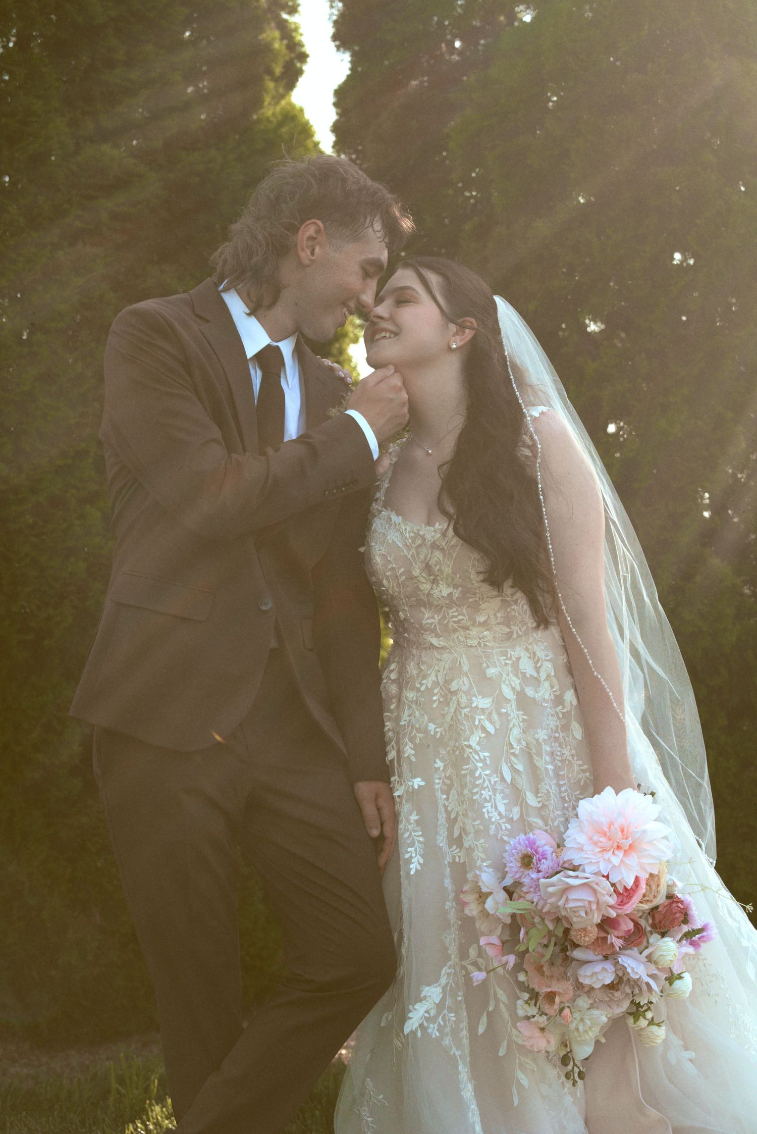 A tender wedding moment captured showing a couple in formal attire sharing a romantic kiss in a sunlit garden setting.