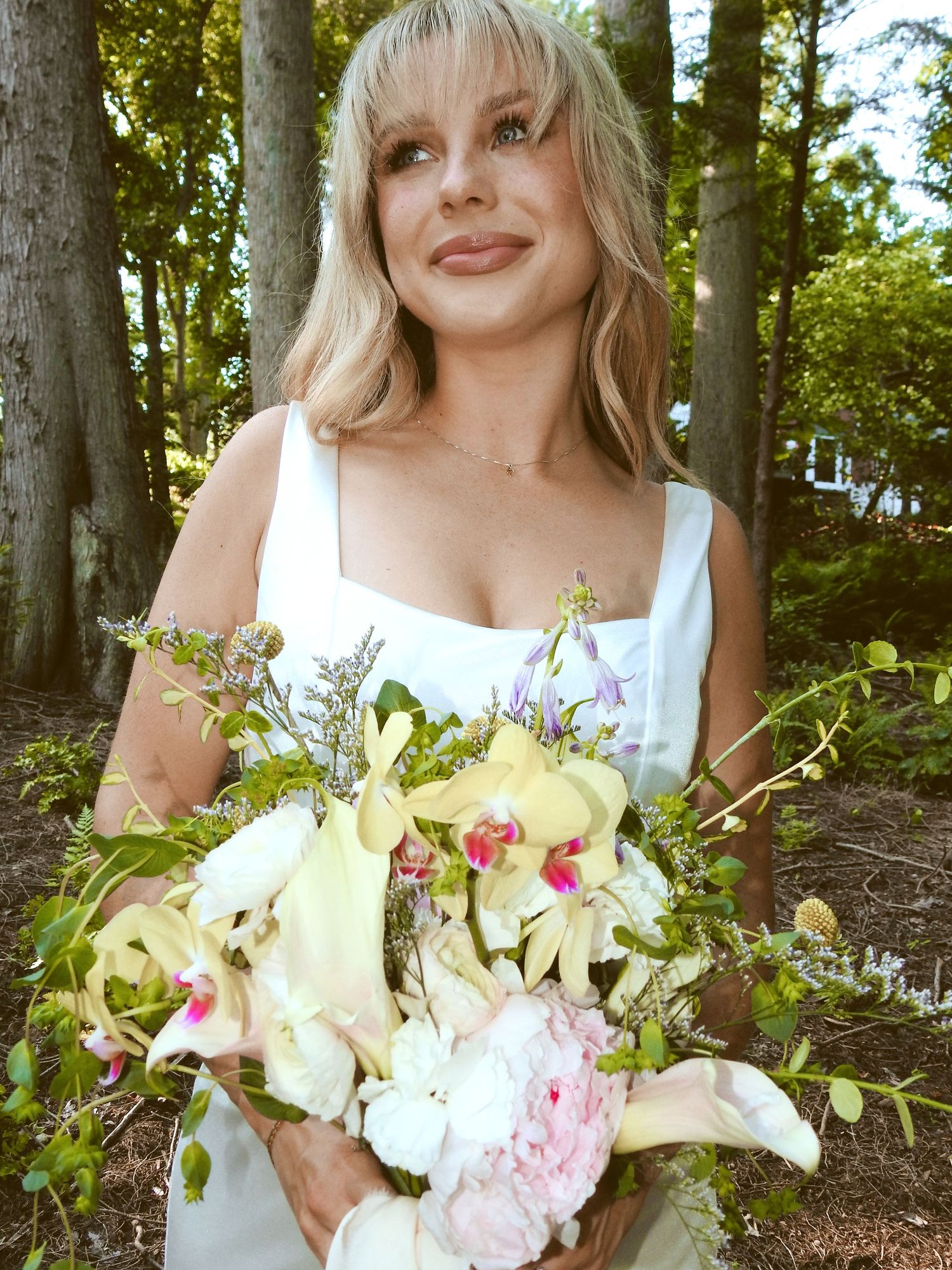 A radiant person in a white dress holds a beautiful bouquet of white lilies, pink peonies and orchids in a forest setting.