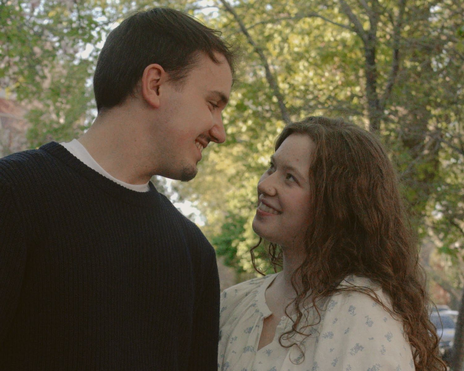 A couple holds hands while walking through a sunlit park with trees in the background.