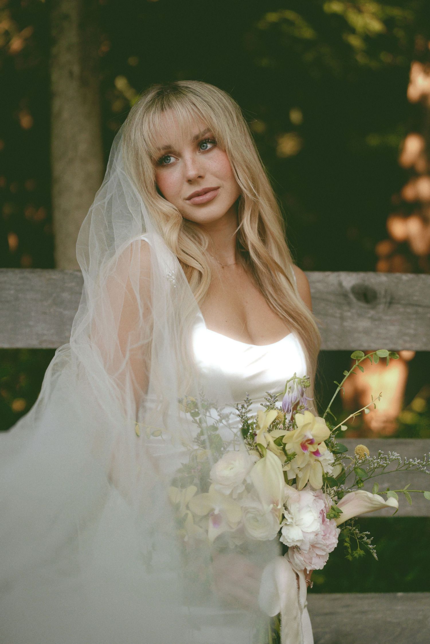 Bride in white wedding dress holding delicate floral bouquet poses against wooden fence in outdoor setting.