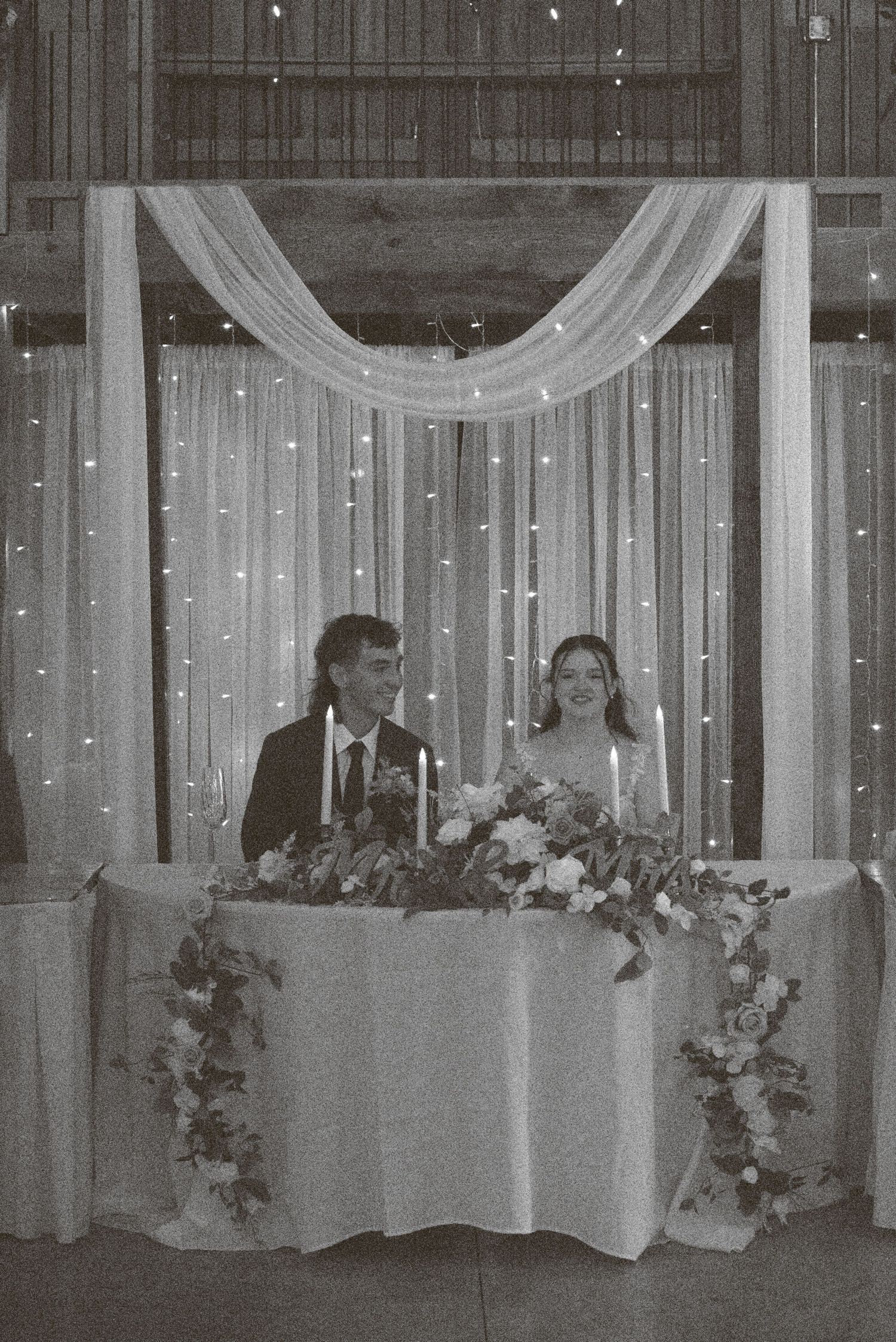 Elegant wedding reception venue with white draped fabric, string lights, and floral arrangements on long dining tables.