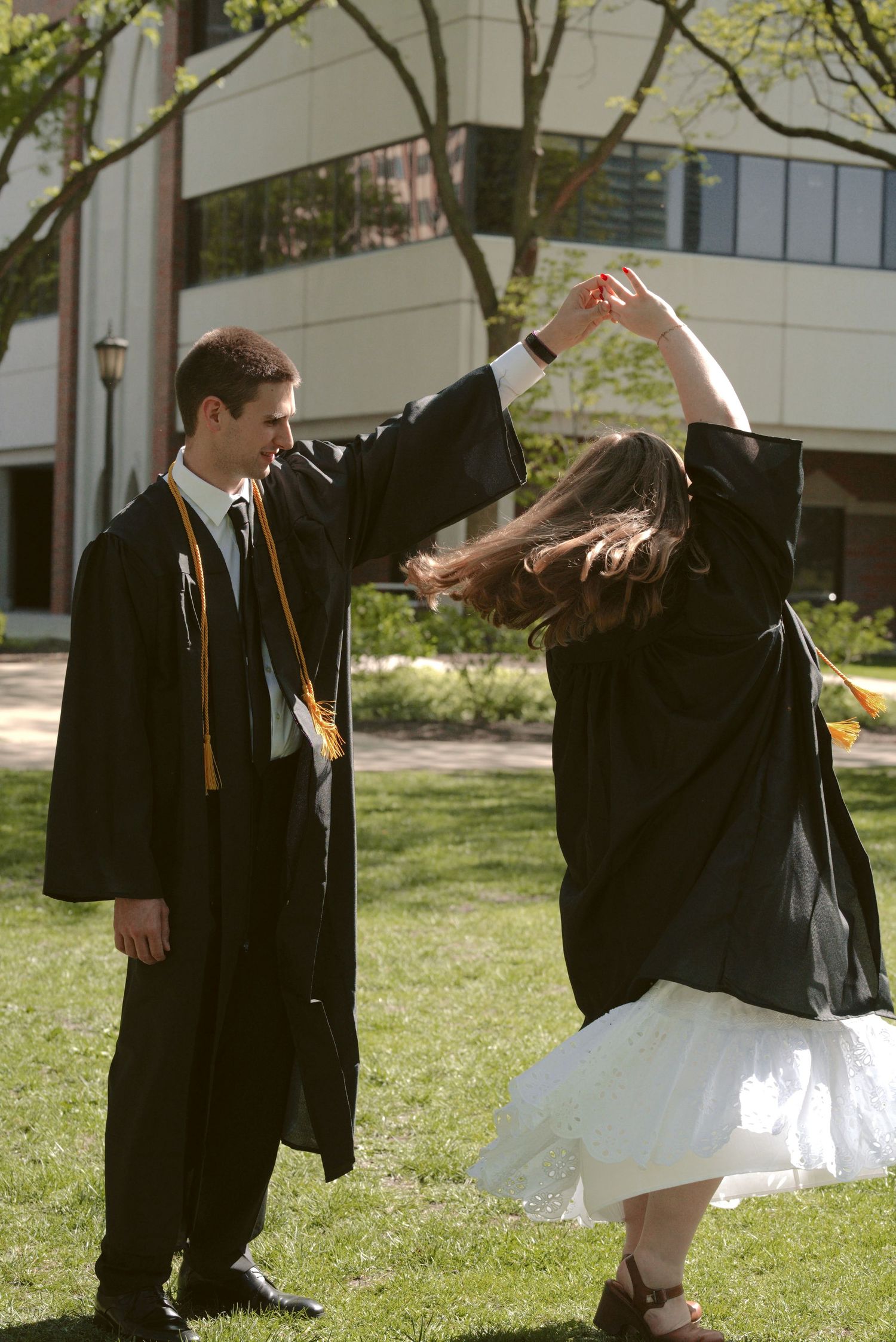 Graduates in black robes dancing and celebrating on sunny campus grounds.