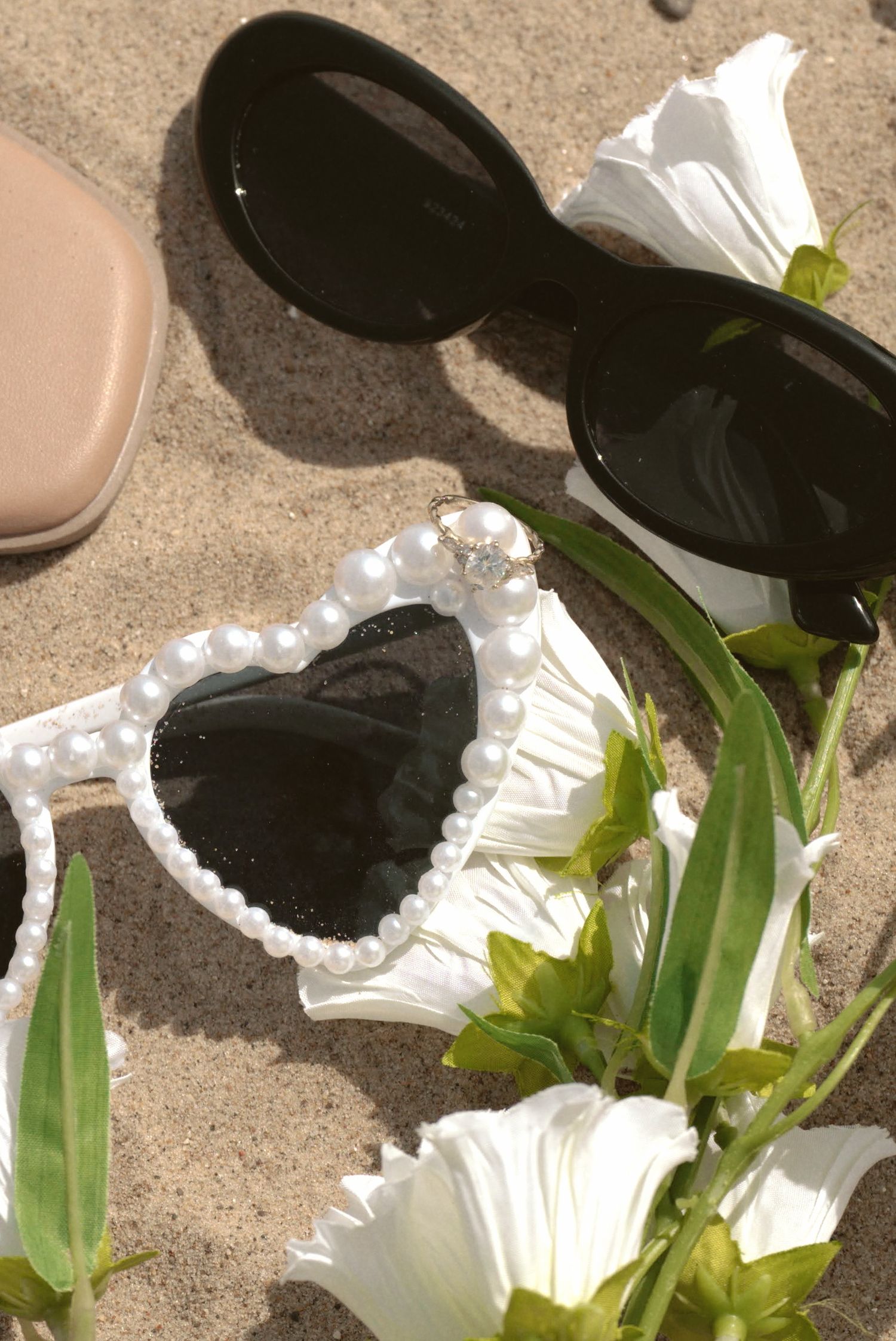Heart-shaped black sunglasses with white floral trim lay on sandy beach next to white petunias and greenery.