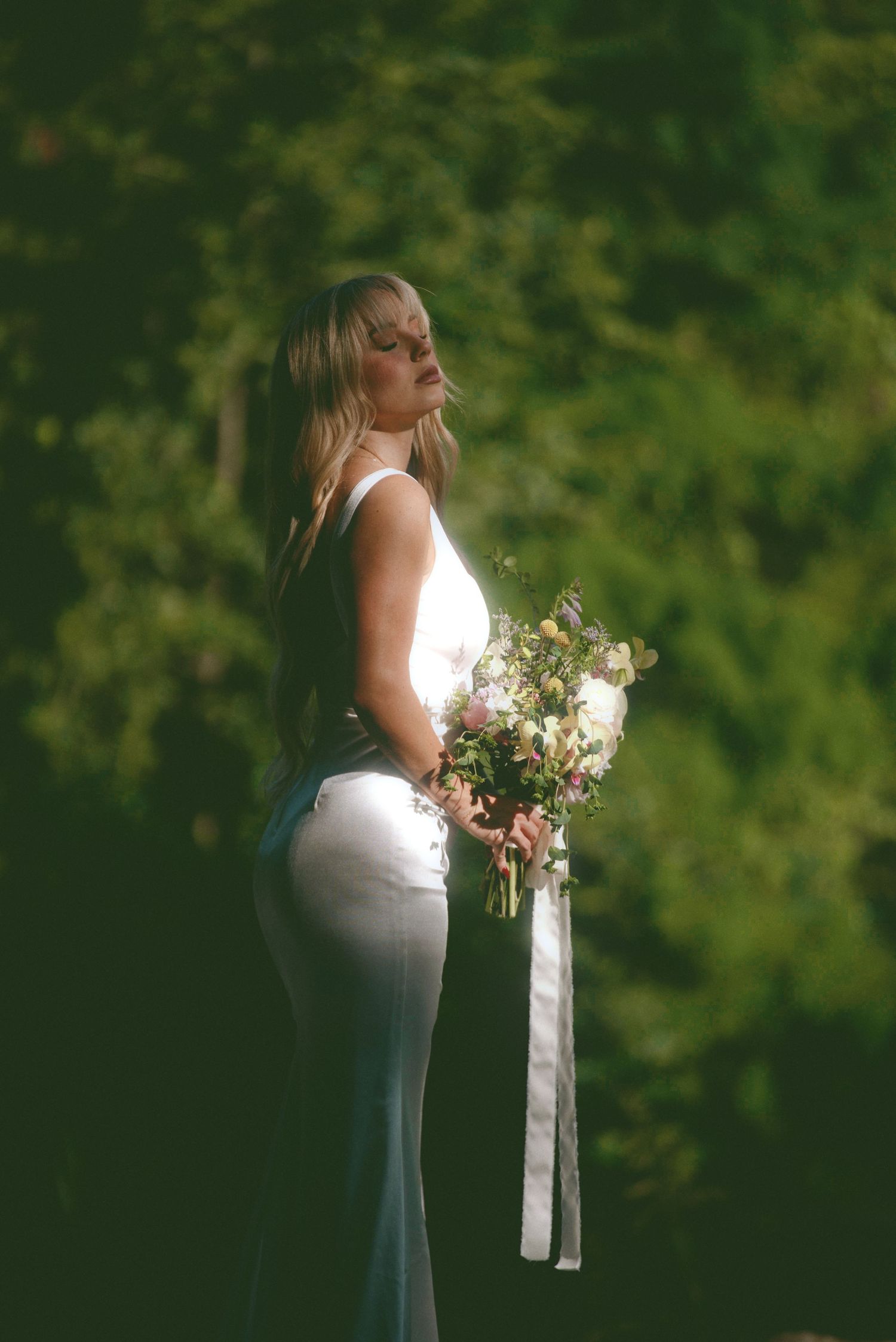 A person in a white wedding dress stands in soft lighting against a green forest backdrop holding a floral bouquet.