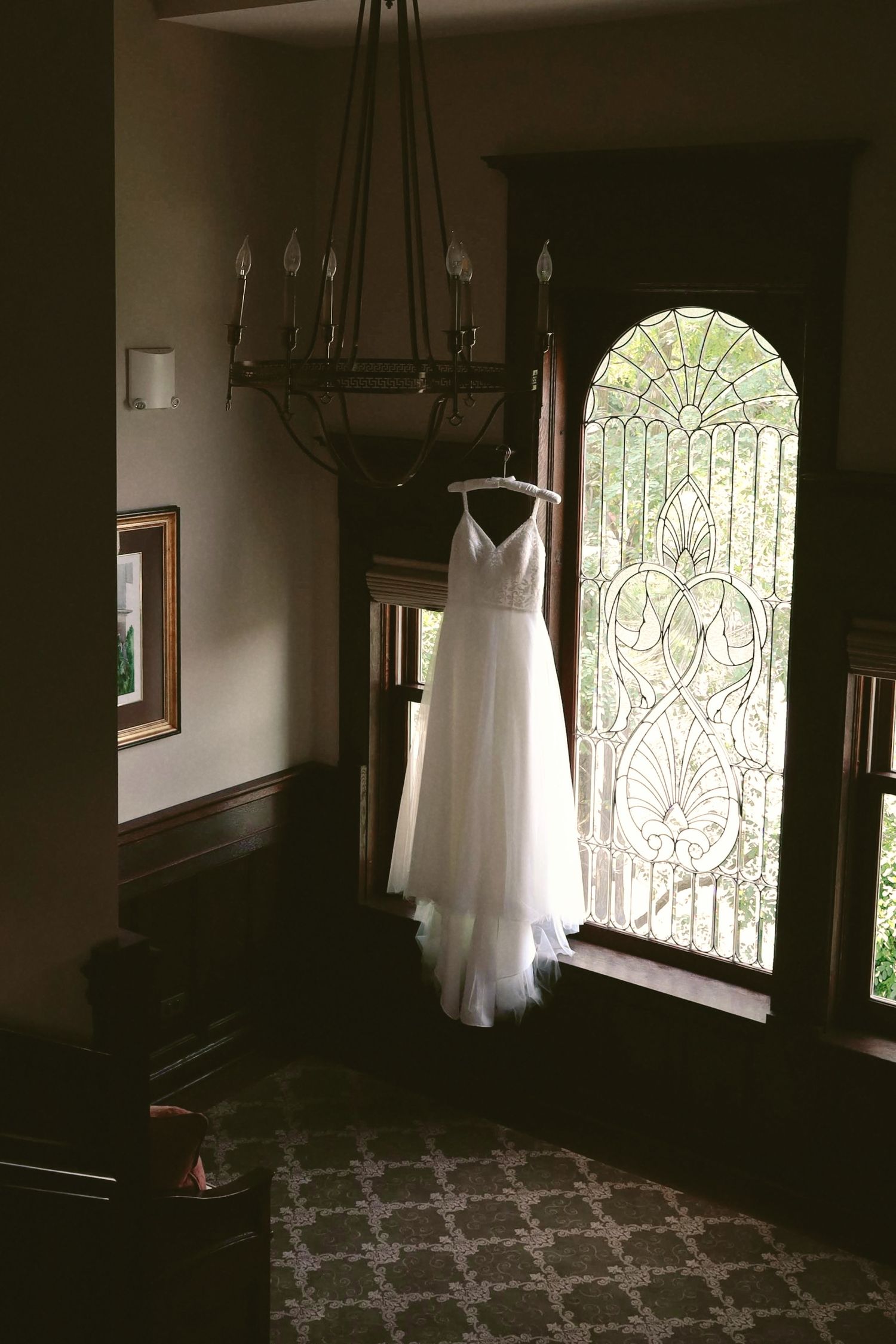 A white wedding dress hangs silhouetted against a bright window in a dark room creating a dramatic lighting effect.