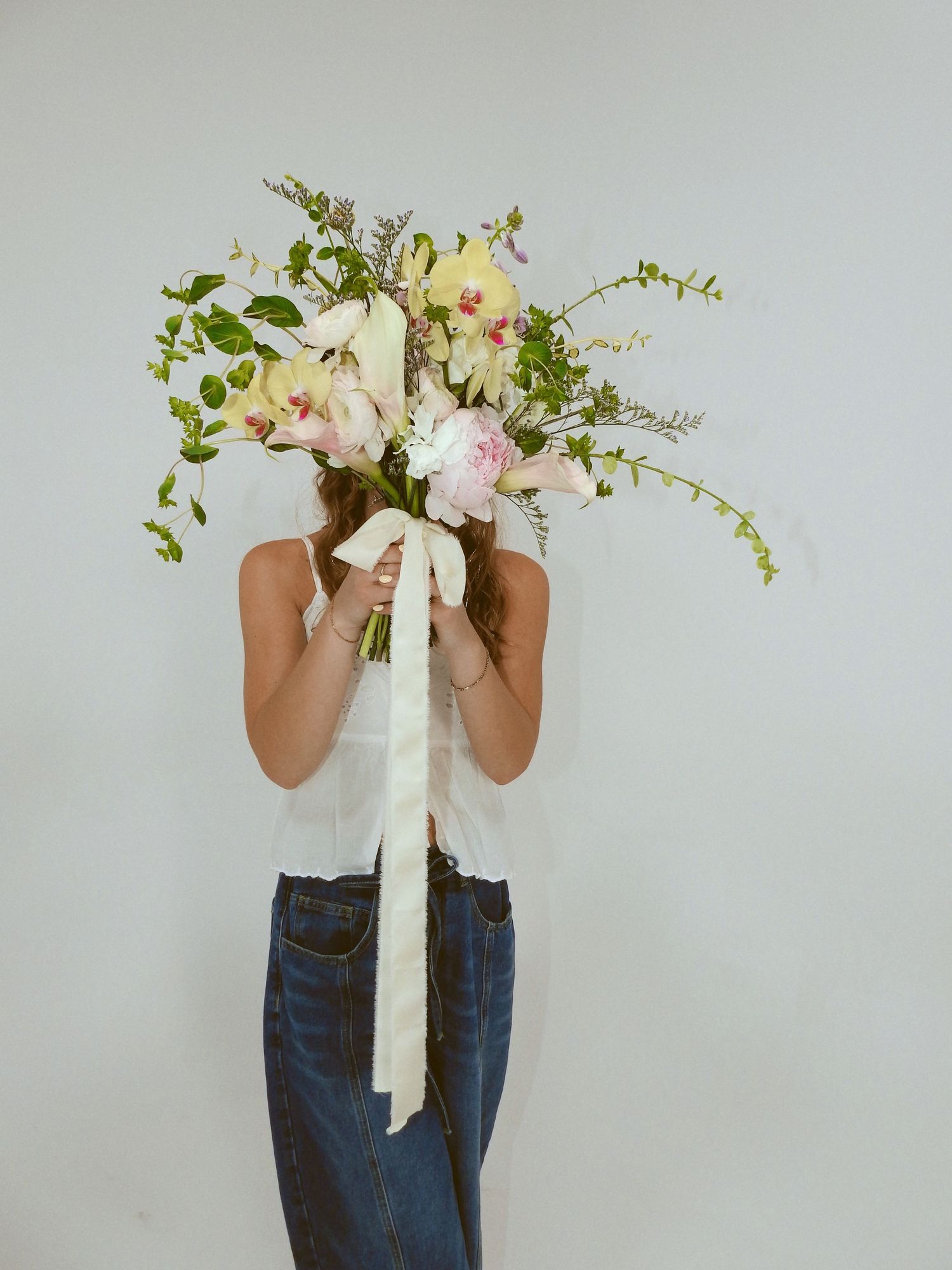 A person wearing blue jeans and white top holds a delicate floral arrangement featuring pink blooms and cascading greenery.