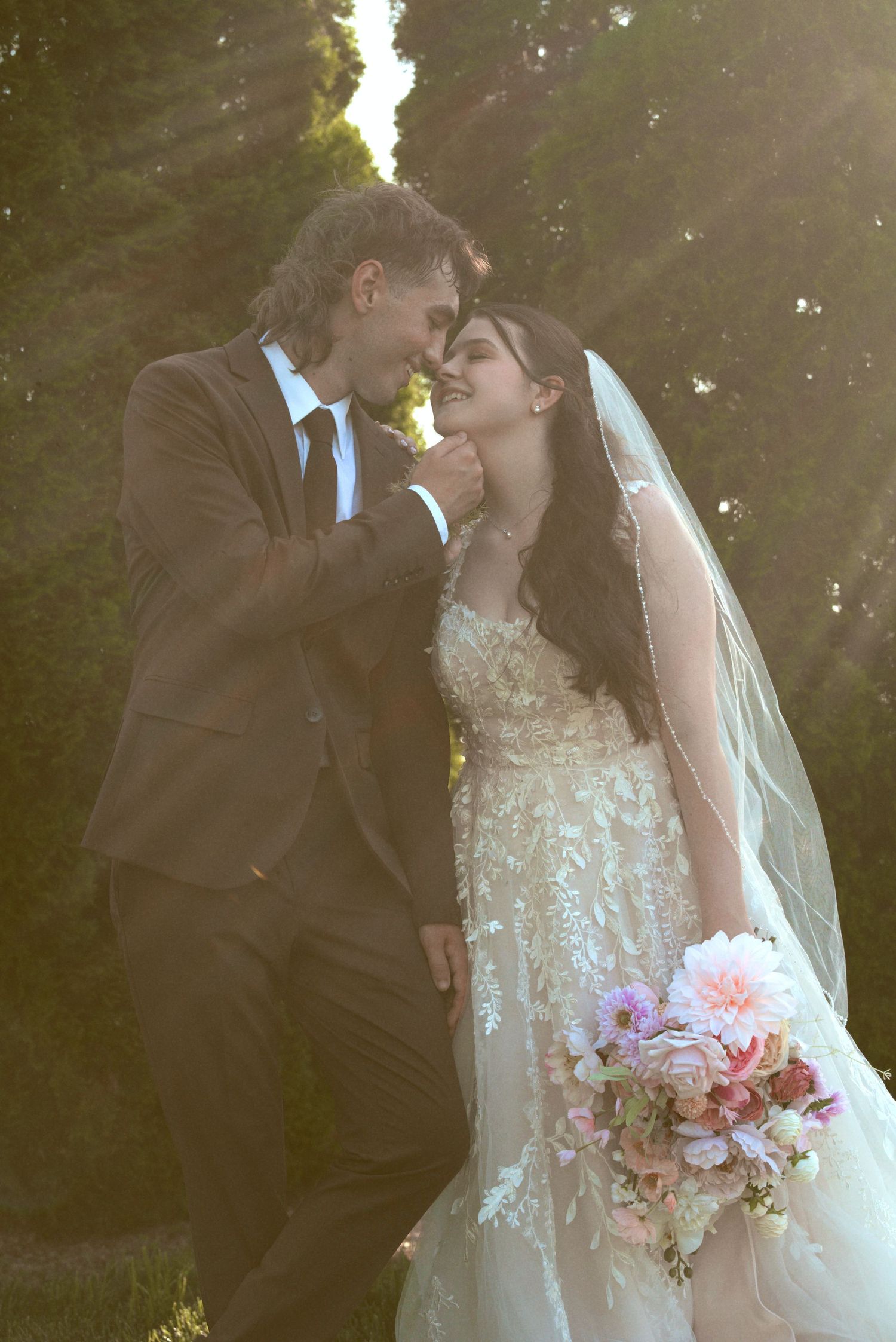 A romantic wedding couple shares an intimate moment at sunset, with the bride holding a pink peony bouquet and wearing a lace gown.
