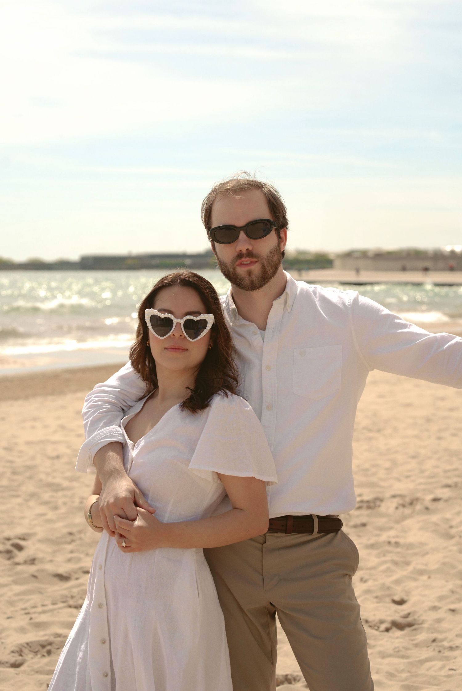 A couple wearing sunglasses embrace on a sunny beach with white sand and ocean waves in the background.