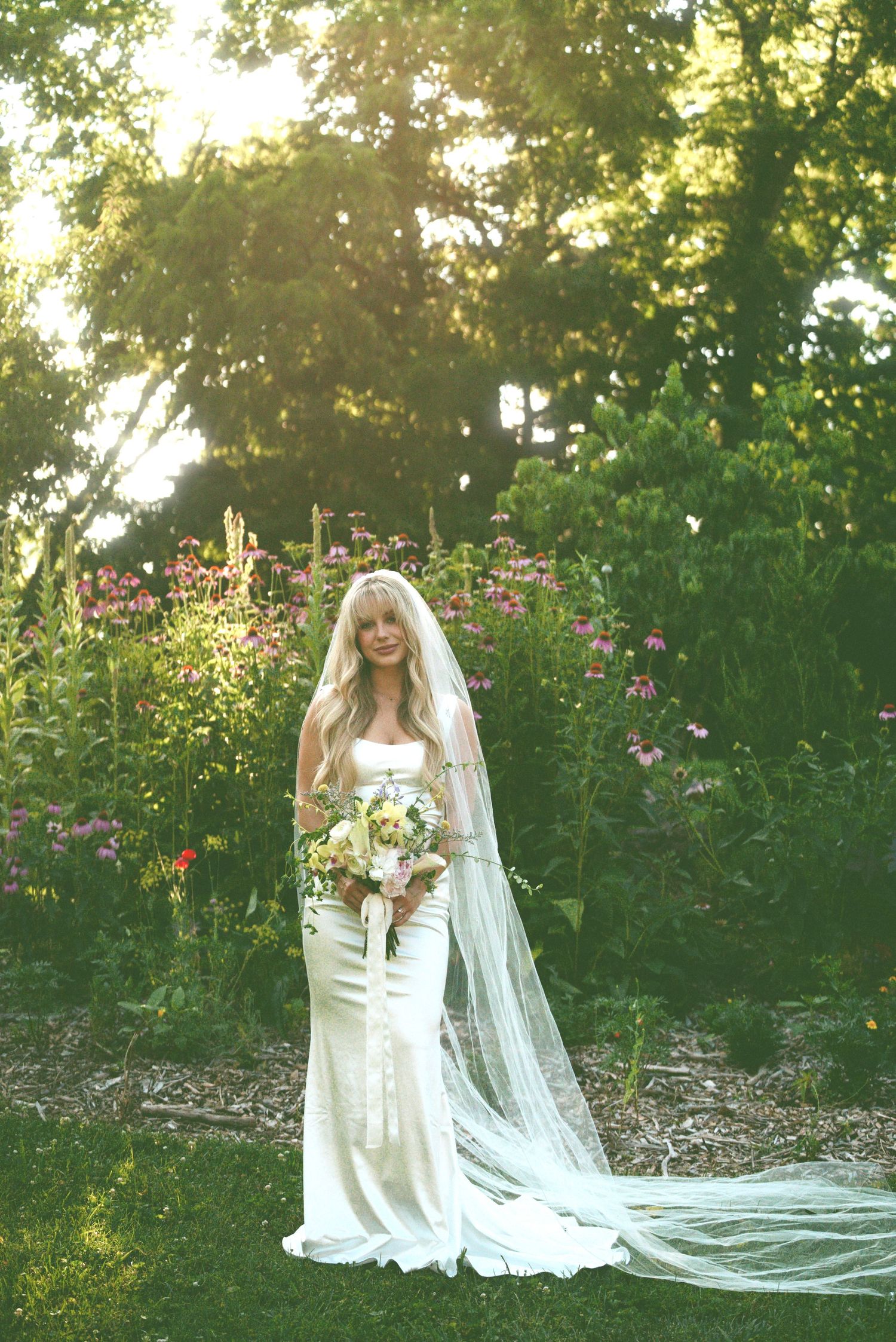 A stunning bride in a flowing lace gown with cathedral veil poses in a lush garden setting during golden hour.