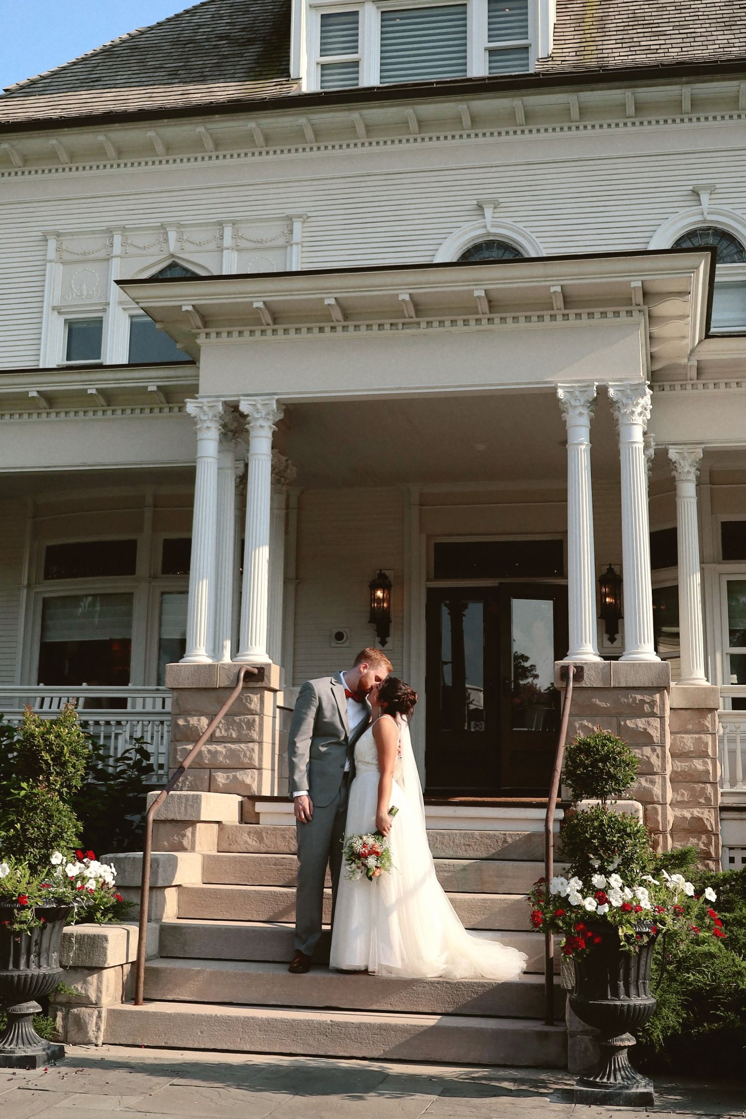 A couple stands on the steps of a grand white Victorian mansion with ornate columns and flower decorations.