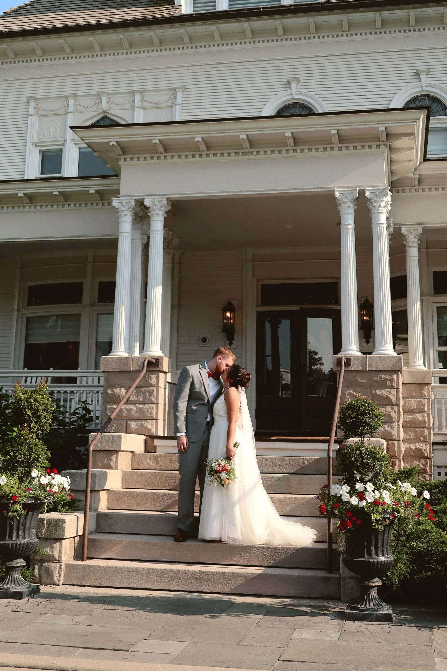 A couple stands on the stone steps of a grand white Victorian mansion with decorative columns and potted flowers.