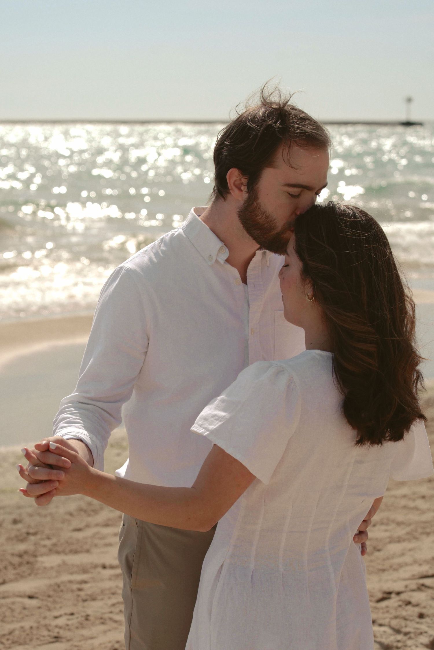 A romantic couple in white clothes dancing together on a sunny beach at sunset.
