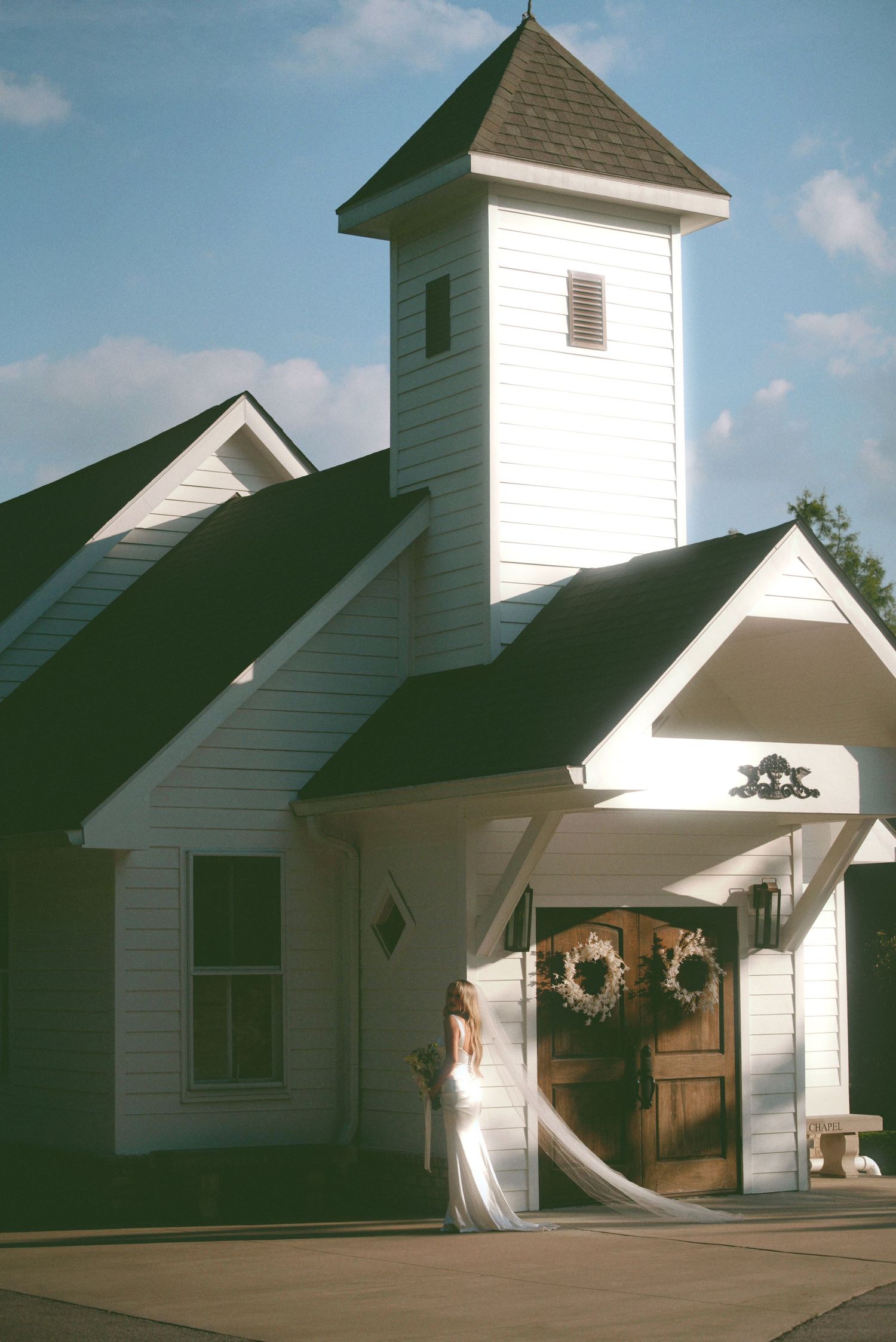 A white country church with a tall steeple tower and green roof features a festive wreath on its wooden front door.