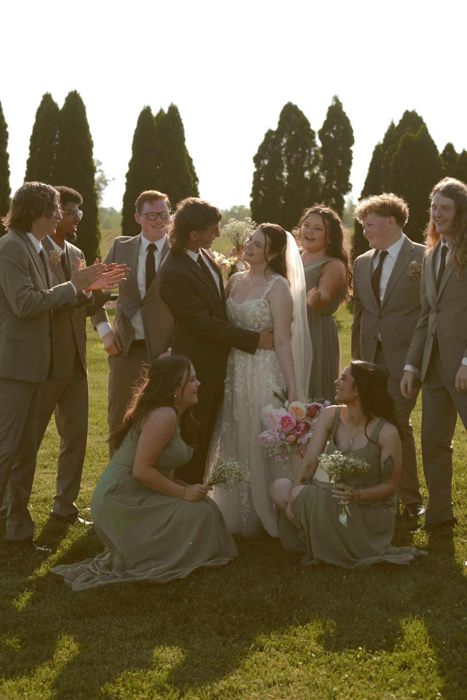 Wedding party with bridesmaids in sage green dresses and groomsmen in gray suits celebrate in a sunlit garden.