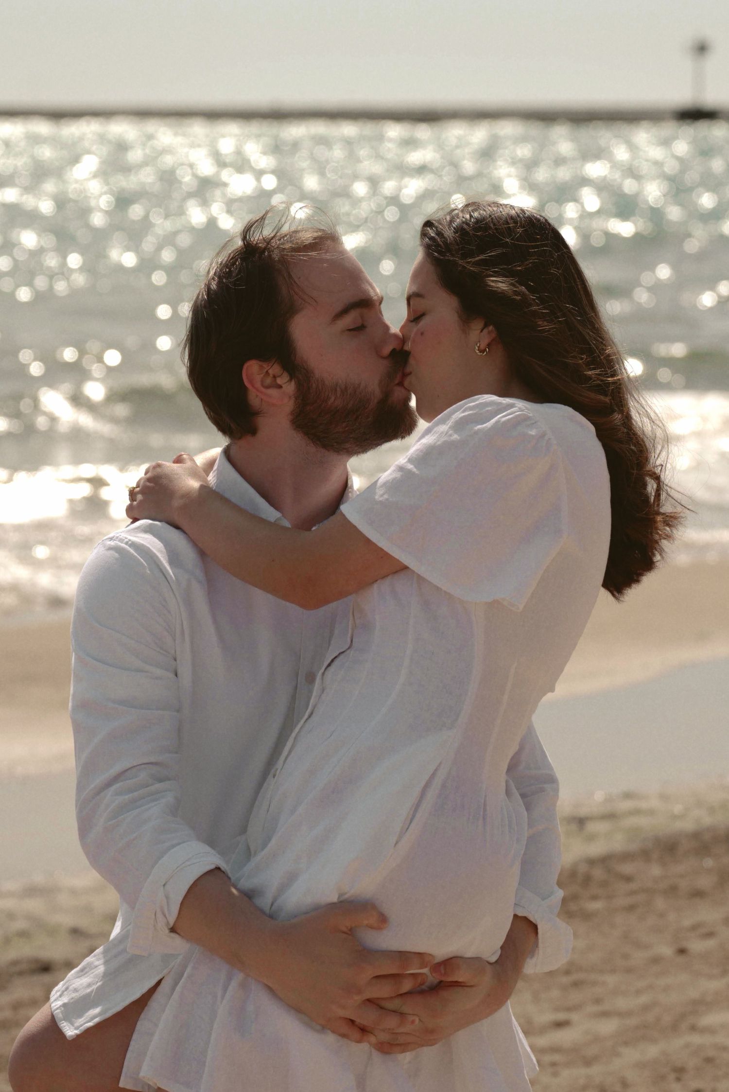 A romantic couple in white clothing shares an intimate embrace and kiss on a sunlit beach.