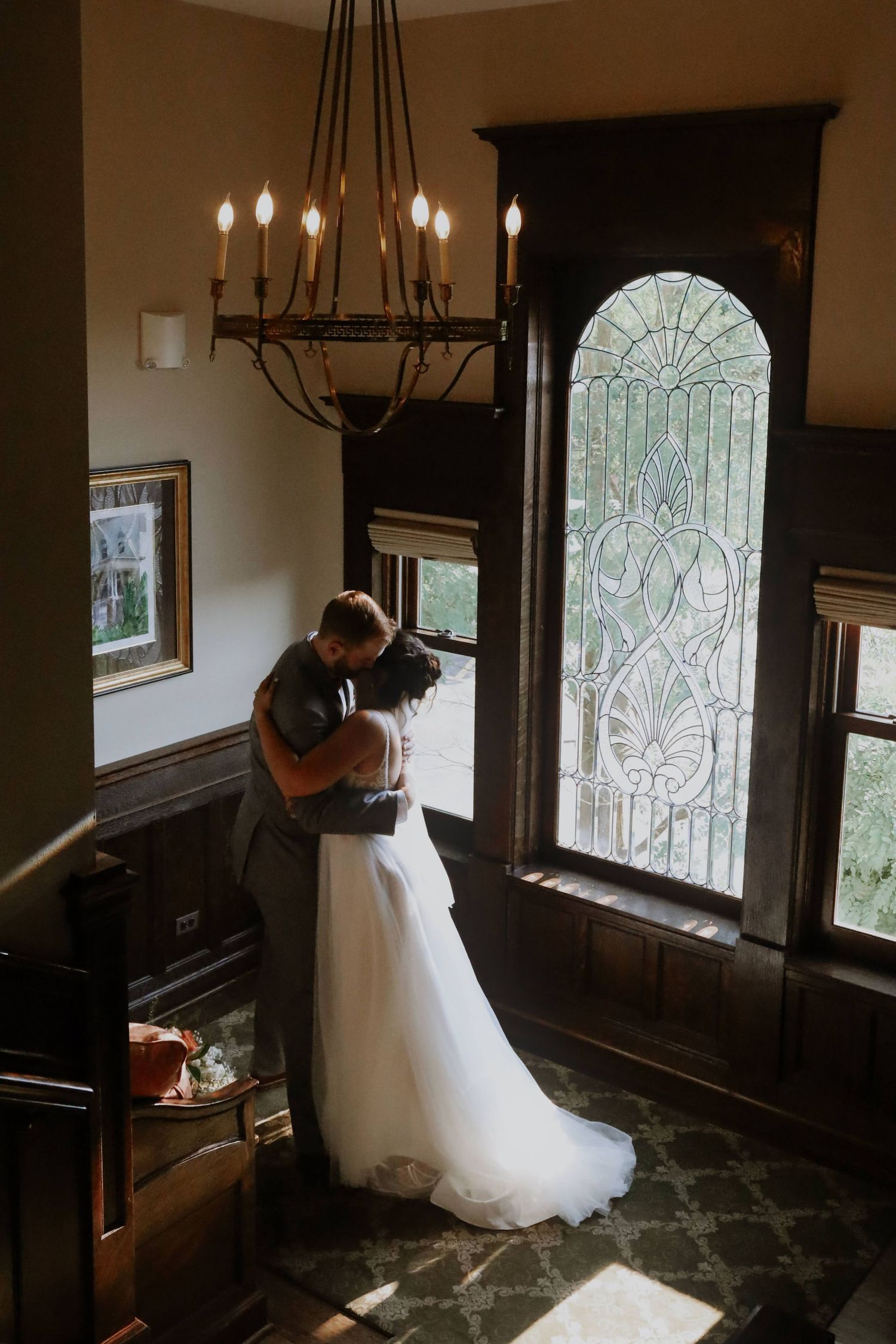 A tender moment shared in front of a stunning stained glass window while a vintage chandelier hangs above in an elegant room.