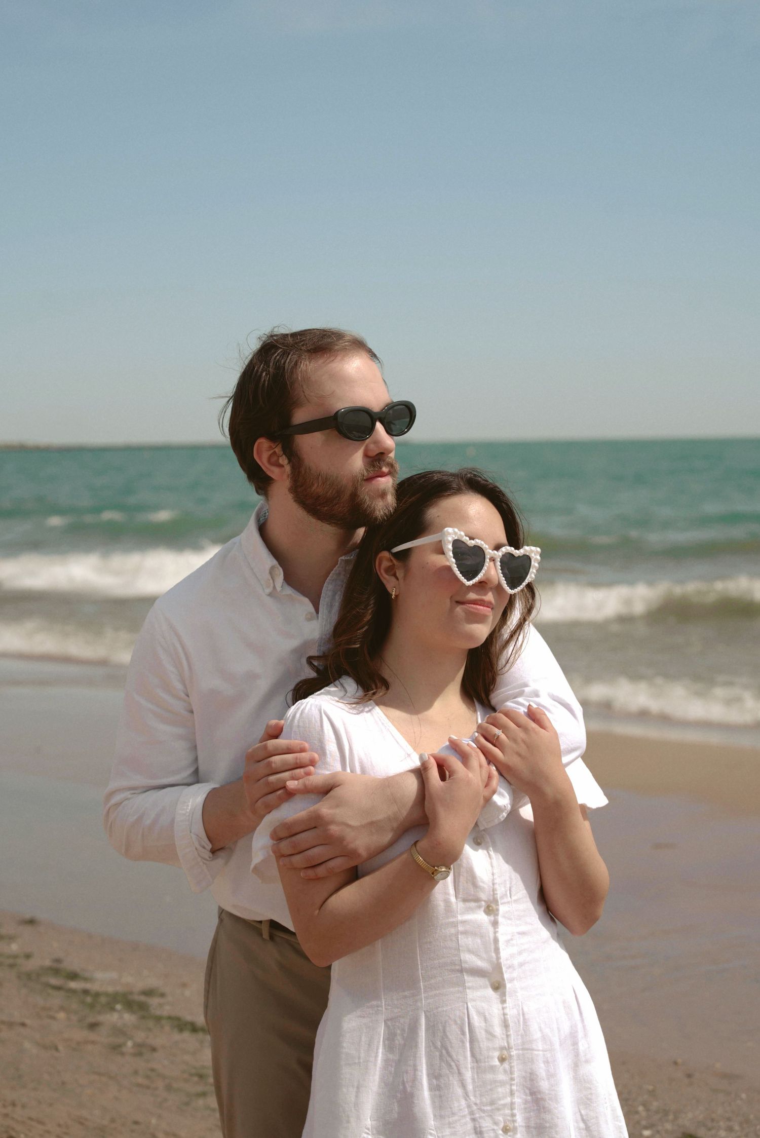 A romantic couple embraces on a sunny beach, wearing sunglasses and light colored clothing by the ocean.
